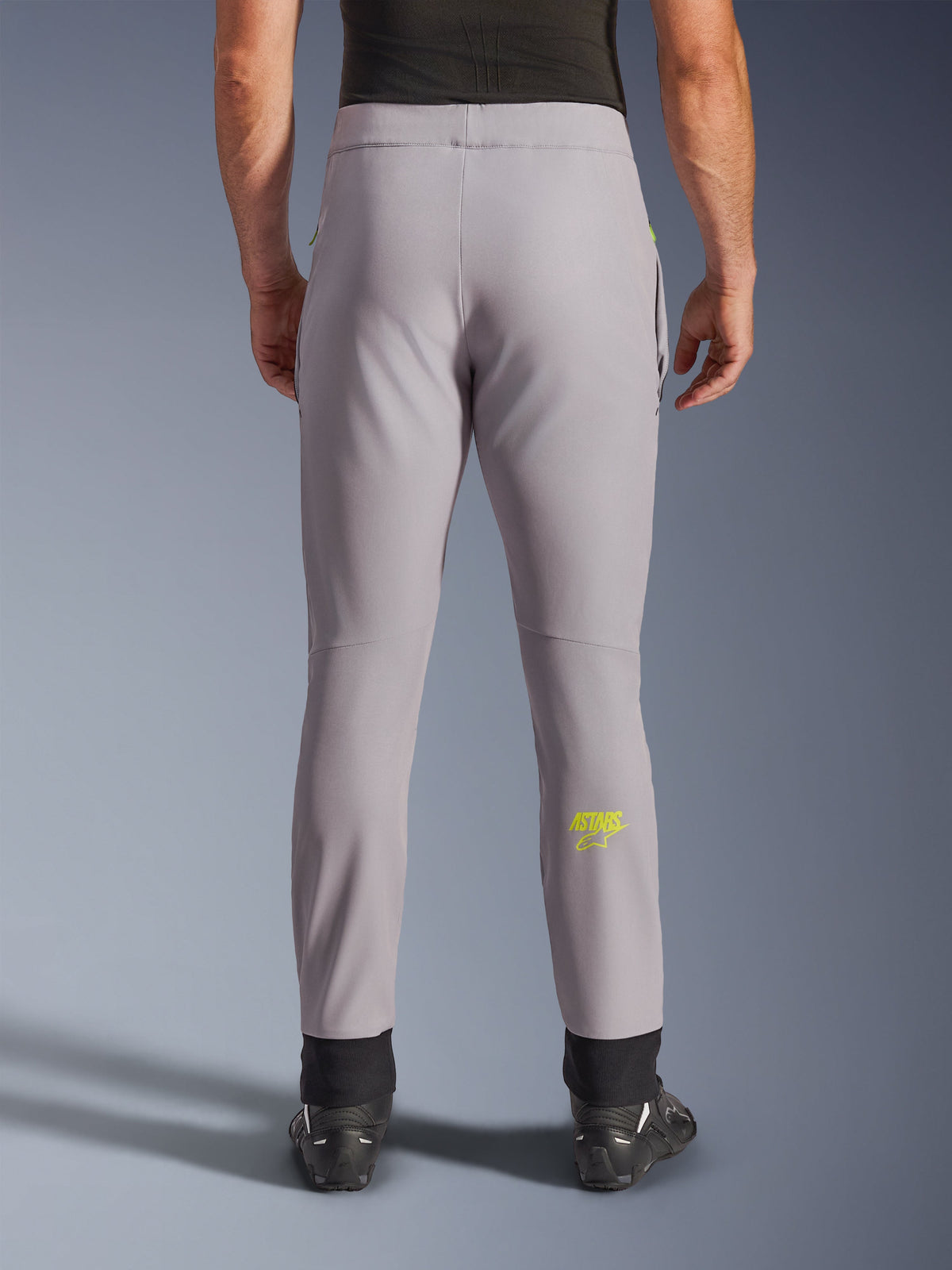 Alpinestars Aeroshell Pants, spodnie motocyklowe, szare i żółte neonowe, widok z tyłu przedstawiający żółte logo Astars na łydce, czarne elastyczne ściągacze na kostkach, nowoczesny design softshell do jazdy szosowej i codziennej