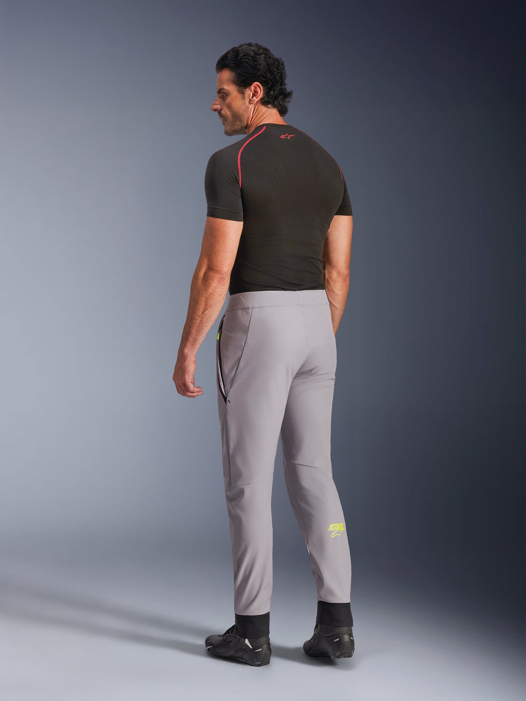 Spodnie Alpinestars Aeroshell, Mid Gray Yellow Fluo, widok z tyłu na modela w elastycznych motocyklowych spodniach softshellowych, żółte logo Astars na lewej łydce, w zestawieniu z czarną warstwą bazową i butami motocyklowymi, nowoczesna sportowa sylwetka.
