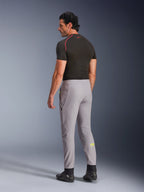 Spodnie Alpinestars Aeroshell, Mid Gray Yellow Fluo, widok z tyłu na modela w elastycznych motocyklowych spodniach softshellowych, żółte logo Astars na lewej łydce, w zestawieniu z czarną warstwą bazową i butami motocyklowymi, nowoczesna sportowa sylwetka.