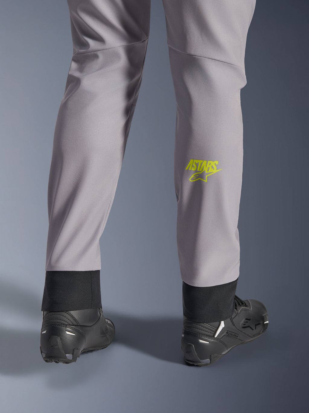 Alpinestars Aeroshell Pants, średnioszare z żółtym fluo logo Astars na łydce, widok od tyłu w czarnych butach motocyklowych, elastyczny dolny brzeg i nowoczesna zwężana sylwetka