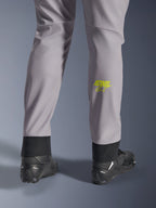Alpinestars Aeroshell Pants, średnioszare z żółtym fluo logo Astars na łydce, widok od tyłu w czarnych butach motocyklowych, elastyczny dolny brzeg i nowoczesna zwężana sylwetka