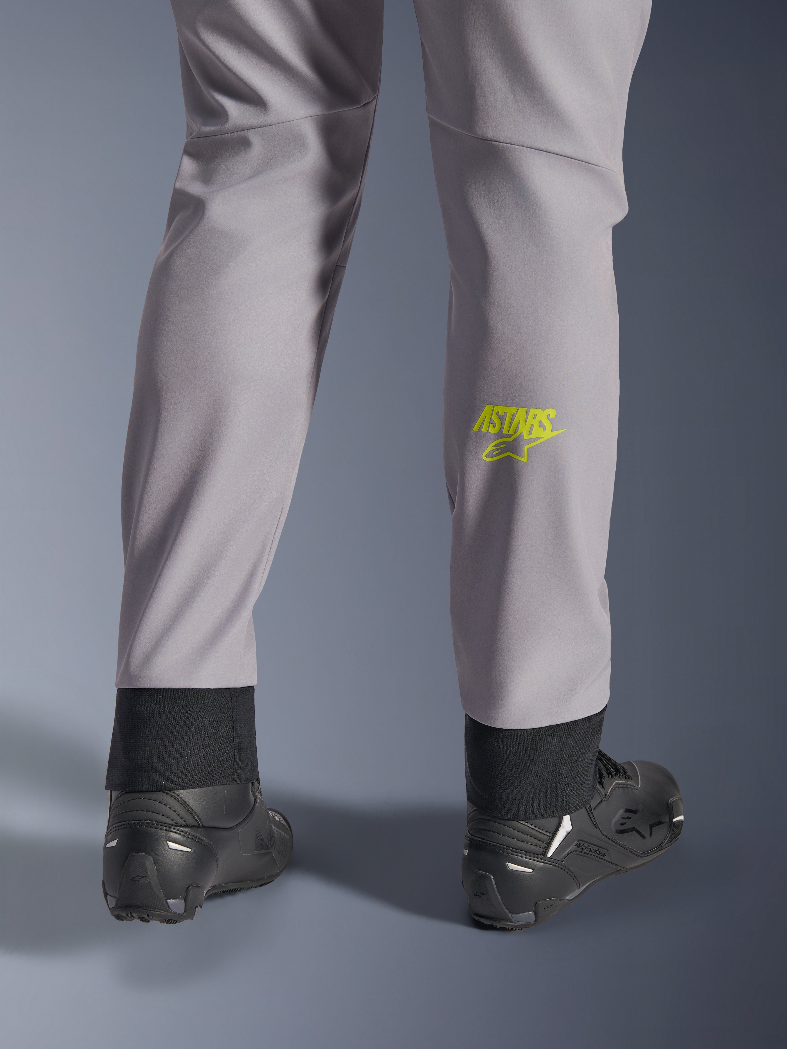 Alpinestars Aeroshell Pants, średnioszare z żółtym fluo logo Astars na łydce, widok od tyłu w czarnych butach motocyklowych, elastyczny dolny brzeg i nowoczesna zwężana sylwetka