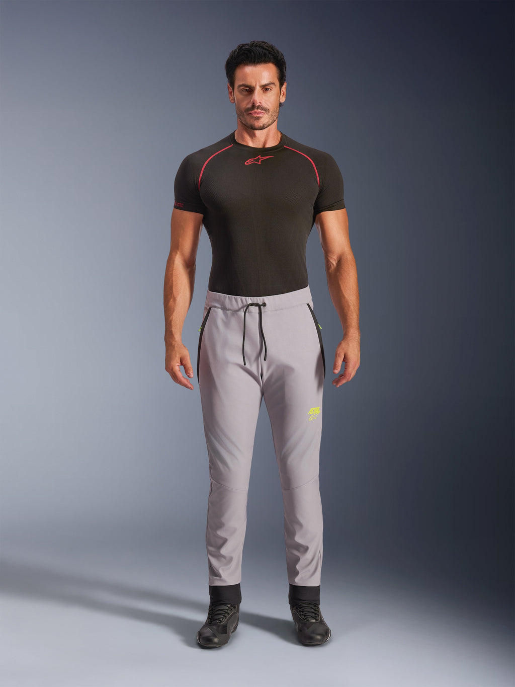 Męski model ubrany w spodnie Alpinestars Aeroshell w kolorze szarym i neonowym żółtym, sportowe spodnie softshellowe typu crossover z elastycznym pasem ze sznurkiem i kieszeniami na zamek, zestawione z czarną koszulką techniczną z krótkim rękawem, nowoczesny sportowy design do jazdy szosowej i codziennej