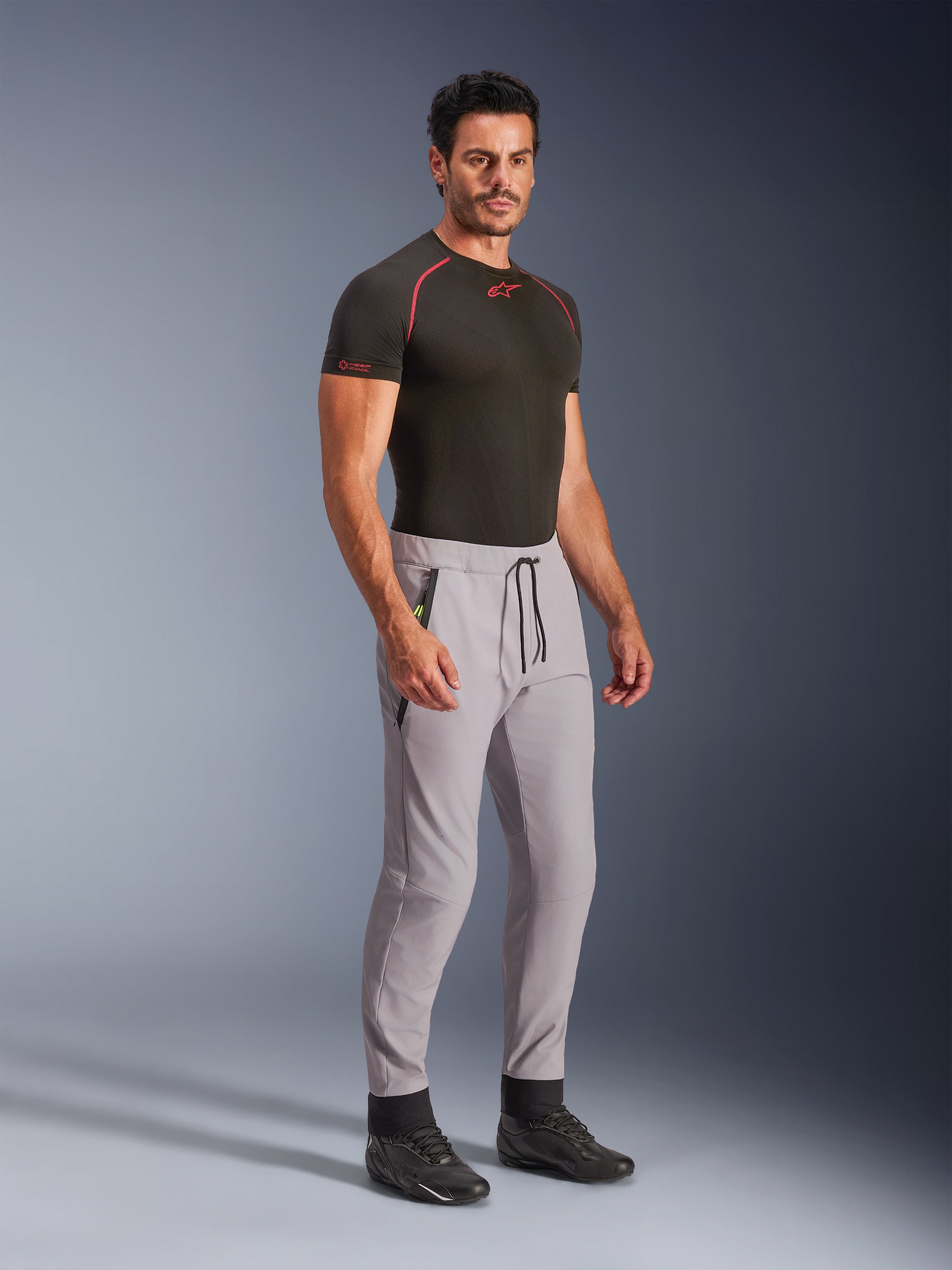 Spodnie Alpinestars Aeroshell, kolor Mid Gray z akcentami Yellow Fluo, sportowe spodnie softshellowe typu crossover prezentowane przez modela w czarnej odzieży termoaktywnej i czarnych butach, nowoczesny zwężany krój, elastyczny pas z czarnym sznurkiem i kieszenie zapinane na zamek.