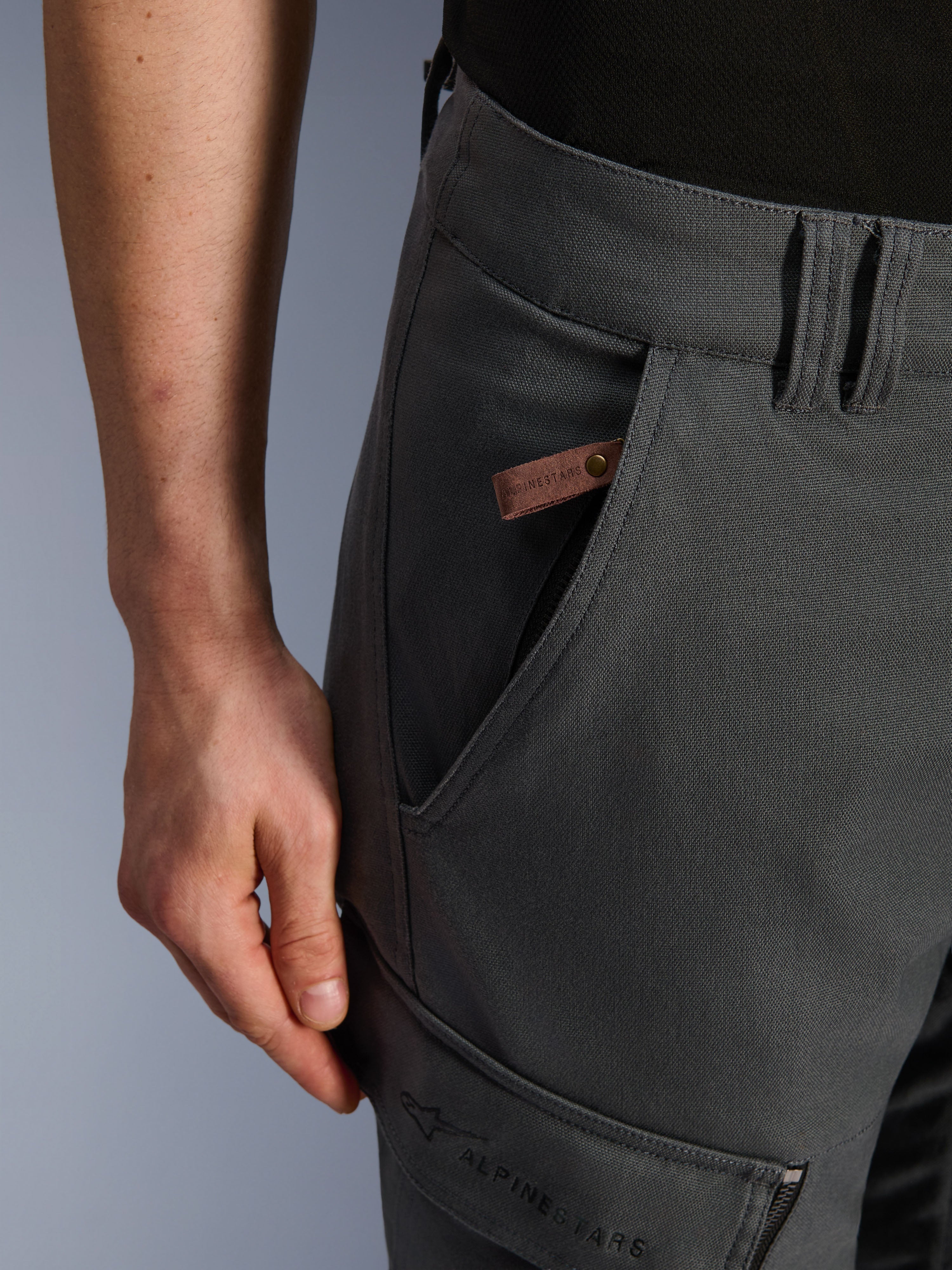 Alpinestars Borrego Drystar® Pants, motocyklowe spodnie adventure, antracytowe, zbliżenie na detal kieszeni i biodra, tekstura woskowanego płótna, brązowa skórzana metka z logo i haftowane oznaczenie Alpinestars na kieszeni cargo na udzie.
