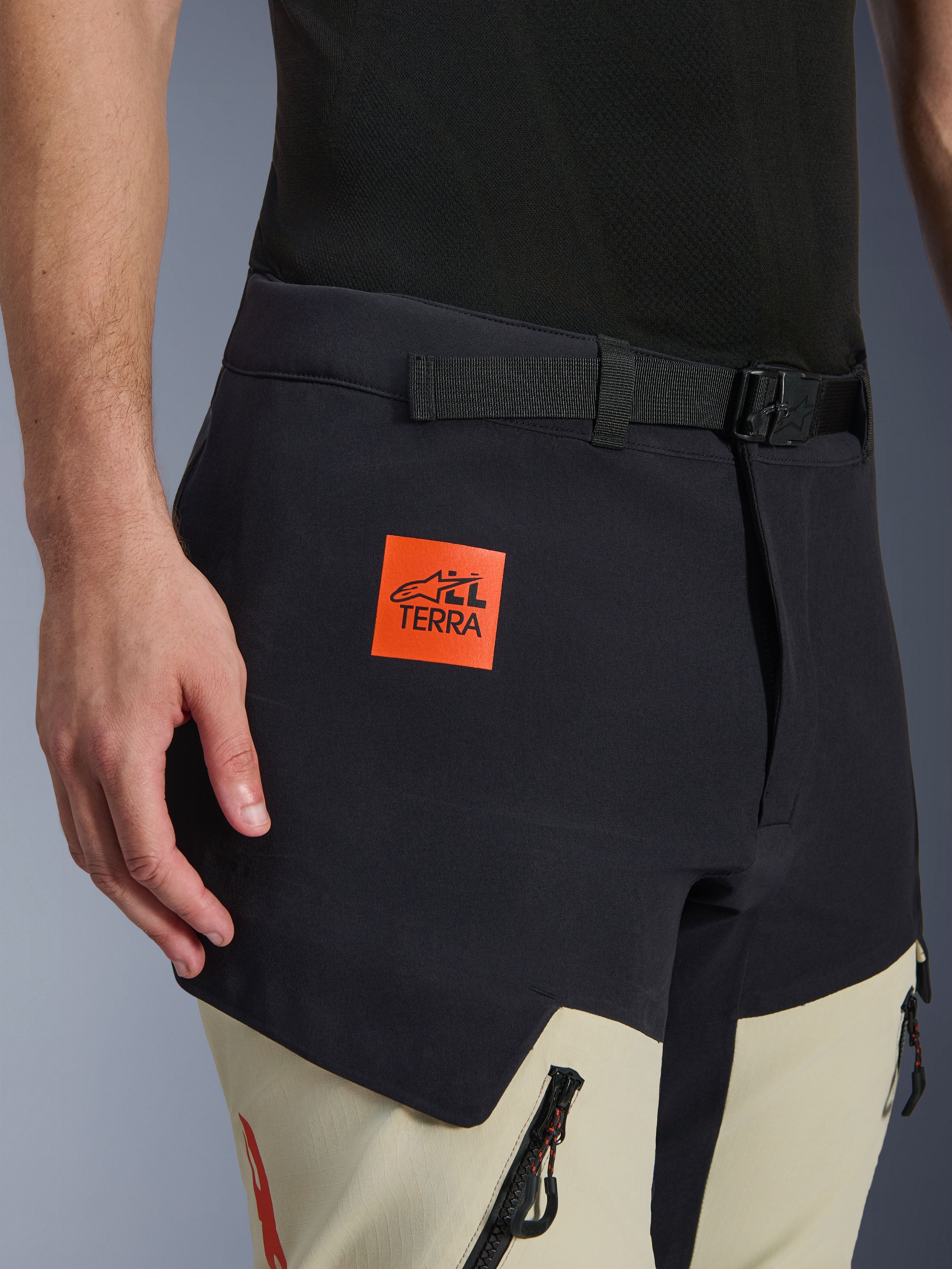 Alpinestars Halo Pro Drystar® XF Laminated Pants, Adventure Motorcycle Pants, Mastic Black and beige, close-up showing the orange 'ALL TERRA' logo patch, black adjustable waist belt, and durable technical fabric with waterproof zippers.