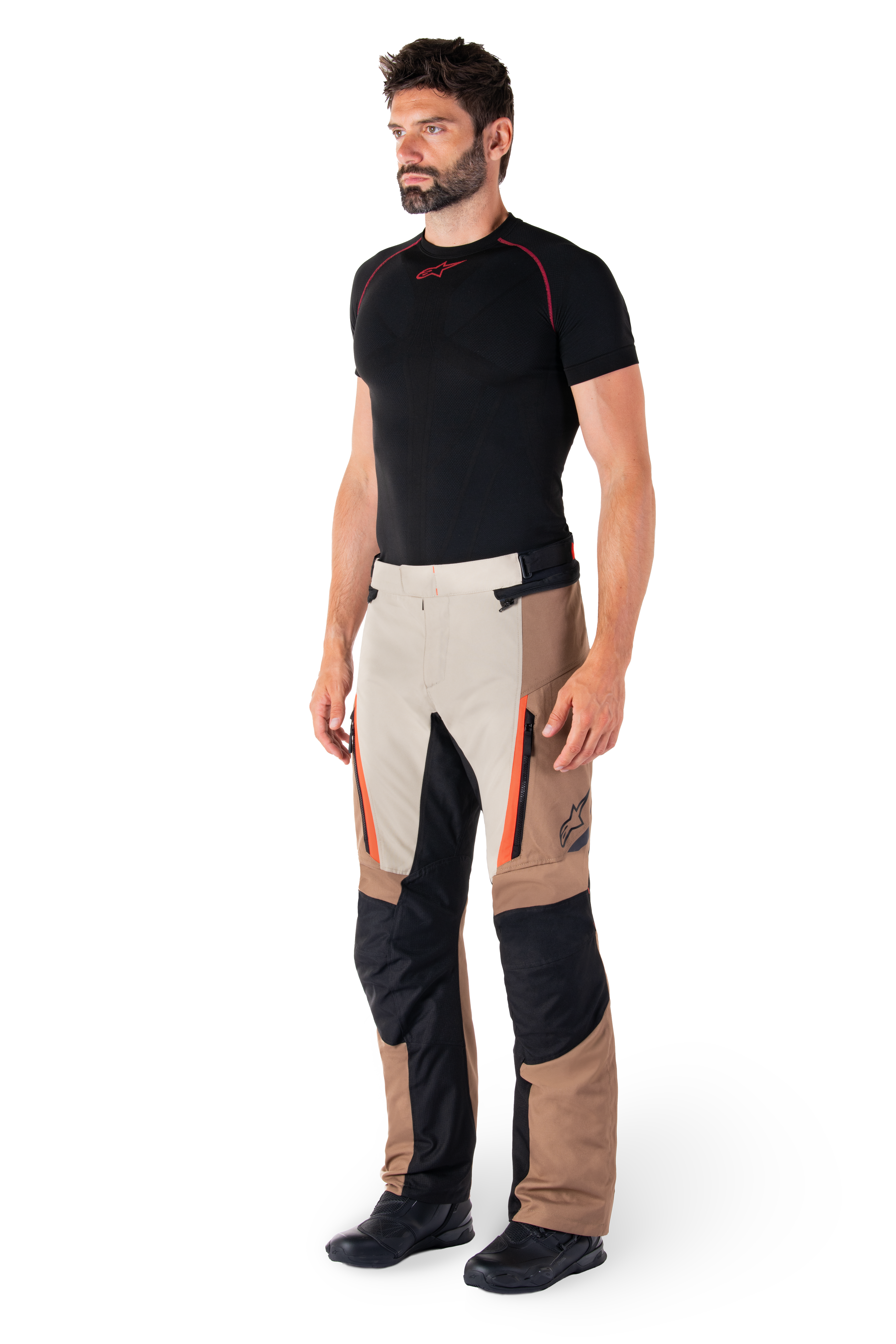 Man wearing Alpinestars ST-1 Waterproof Pants, Adventure Touring Pants, Dark Khaki Sand Black and Red Fluo, styled with a black base layer shirt and black motorcycle boots, featuring reinforced panels and orange zipper accents