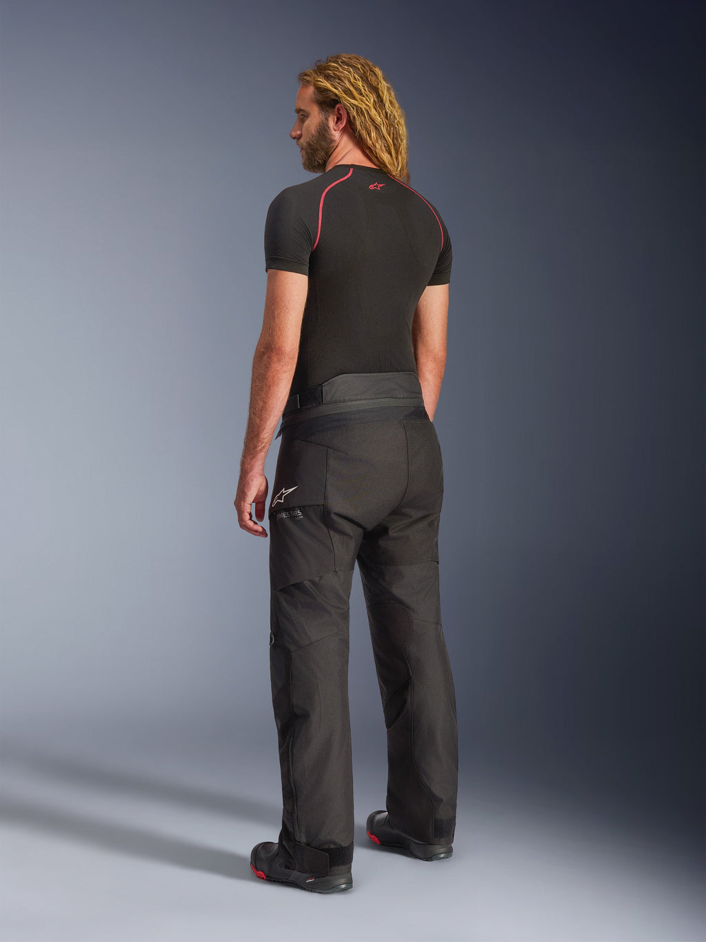 Spodnie Alpinestars Andes V4 Drystar® - Long, przygodowe spodnie motocyklowe, czarne, widok z tyłu pod kątem trzech czwartych na modelu, wytrzymała tekstylna konstrukcja ze wzmocnionymi panelami na siedzeniu i kolanach, z logo gwiazdy Alpinestars, noszone z czarną warstwą termoaktywną i butami motocyklowymi