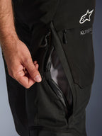 Zbliżenie na czarne spodnie Alpinestars Andes V4 Drystar®, dłoń otwierająca bezpośredni otwór wentylacyjny na udzie z podszewką z siatki, podkreślające wytrzymałą konstrukcję tekstylną i logo Alpinestars.