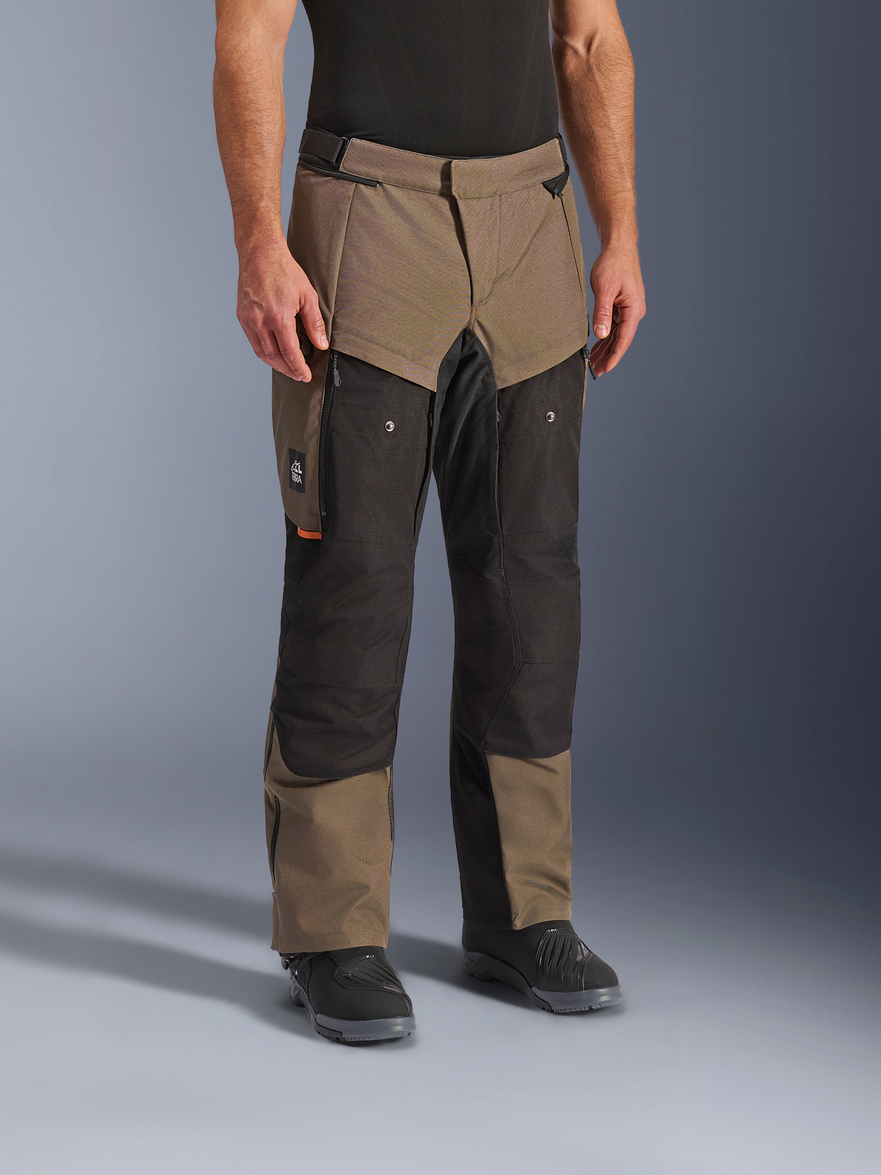 Spodnie Alpinestars Cusco Drystar®, spodnie motocyklowe adventure, Walnut Black, widoczne na modelu w czarnych butach, brązowo-czarna wielopanelowa konstrukcja z kieszeniami na zamek i wzmocnionymi kolanami do turystyki
