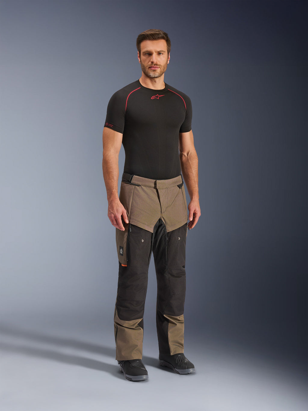 Spodnie Alpinestars Cusco Drystar®, motocyklowe spodnie adventure, w kolorze orzechowym i czarnym, noszone przez modela z czarną warstwą bazową i butami, wyposażone w duże kieszenie cargo na udach, zamki wentylacyjne i wytrzymałą dwukolorową konstrukcję.