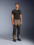 Spodnie Alpinestars Cusco Drystar®, motocyklowe spodnie adventure, w kolorze orzechowym i czarnym, noszone przez modela z czarną warstwą bazową i butami, wyposażone w duże kieszenie cargo na udach, zamki wentylacyjne i wytrzymałą dwukolorową konstrukcję.