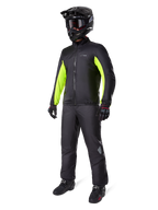 Spodnie Alpinestars Bogota' Pro Drystar® 3 Seasons, czarne, motocyklista w pełnym stroju adventure touring, w tym czarna kurtka z neonowożółtymi akcentami, czarny kask, rękawice i buty, z wodoodporną membraną Drystar® założoną na spodnie.