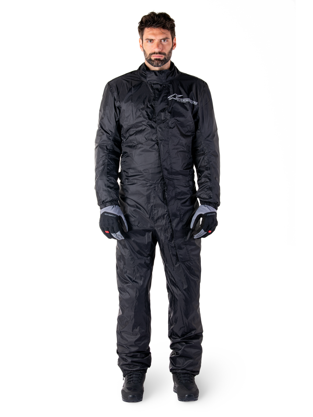 Alpinestars Hurricane Rain V2 Suit, pełny motocyklowy kombinezon przeciwdeszczowy w kolorze czarnym, wodoodporna odzież ochronna z wysokim kołnierzem i przednim zapięciem uszczelniającym, białe logo na piersi, prezentowany z czarno-szarymi rękawicami i butami