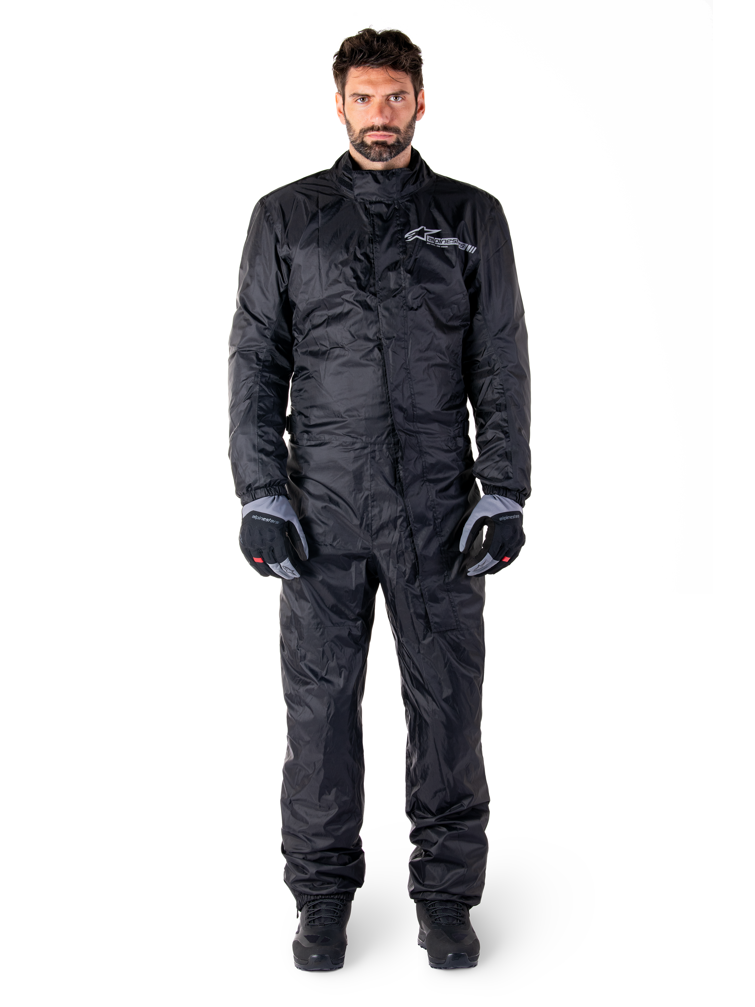 Alpinestars Hurricane Rain V2 Suit, pełny motocyklowy kombinezon przeciwdeszczowy w kolorze czarnym, wodoodporna odzież ochronna z wysokim kołnierzem i przednim zapięciem uszczelniającym, białe logo na piersi, prezentowany z czarno-szarymi rękawicami i butami