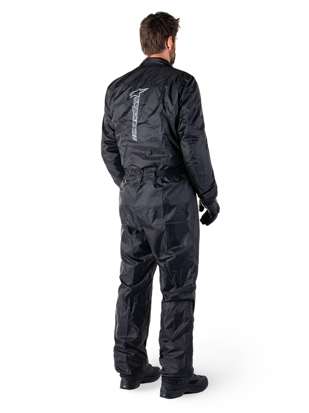 Alpinestars Hurricane Rain V2 Suit, czarny jednoczęściowy wodoodporny motocyklowy kombinezon przeciwdeszczowy, widok z tyłu z dużym pionowym odblaskowym logo, lekka wiatroodporna tkanina, noszony z czarnymi rękawicami i butami