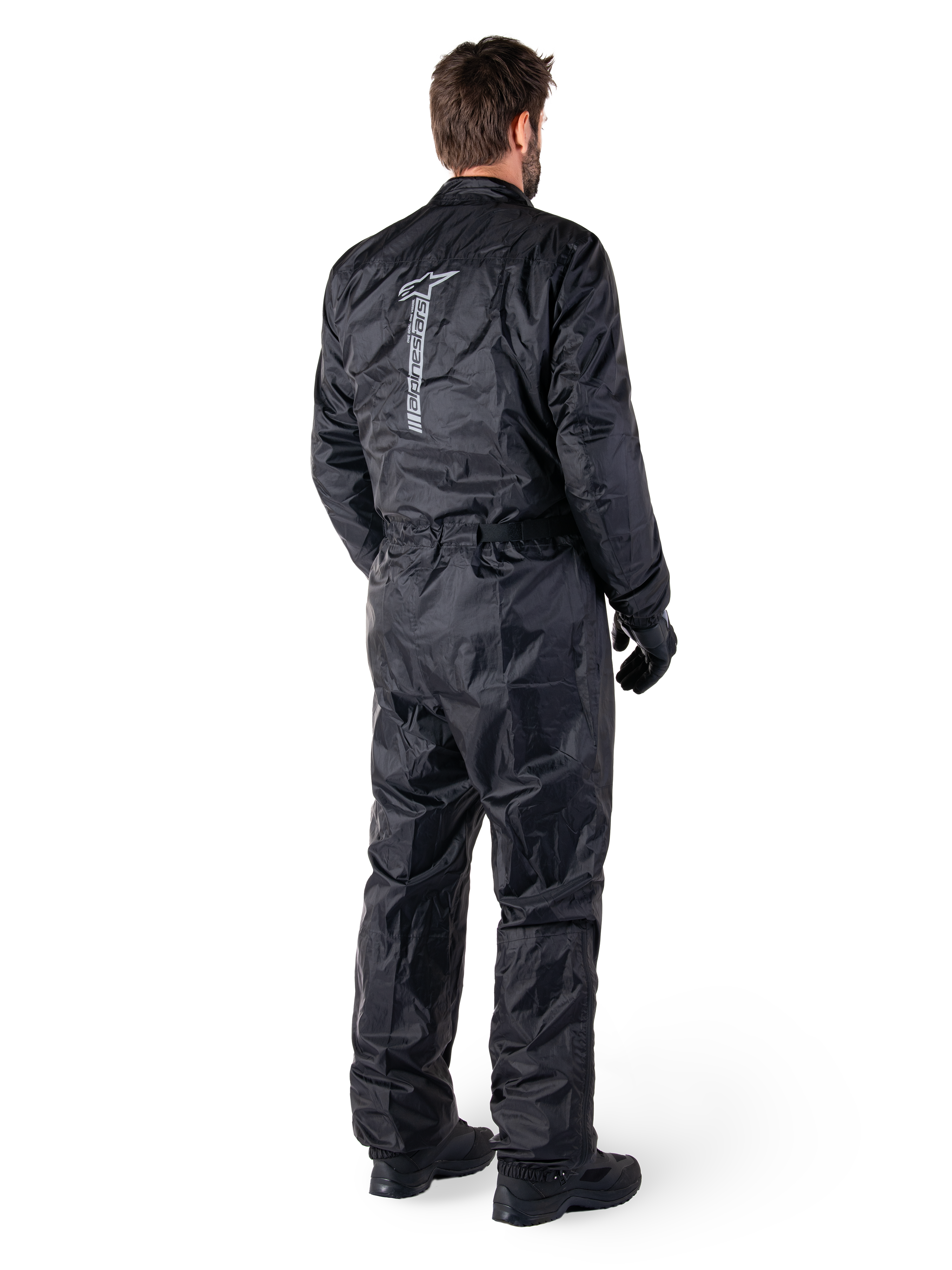 Alpinestars Hurricane Rain V2 Suit, czarny jednoczęściowy wodoodporny motocyklowy kombinezon przeciwdeszczowy, widok z tyłu z dużym pionowym odblaskowym logo, lekka wiatroodporna tkanina, noszony z czarnymi rękawicami i butami