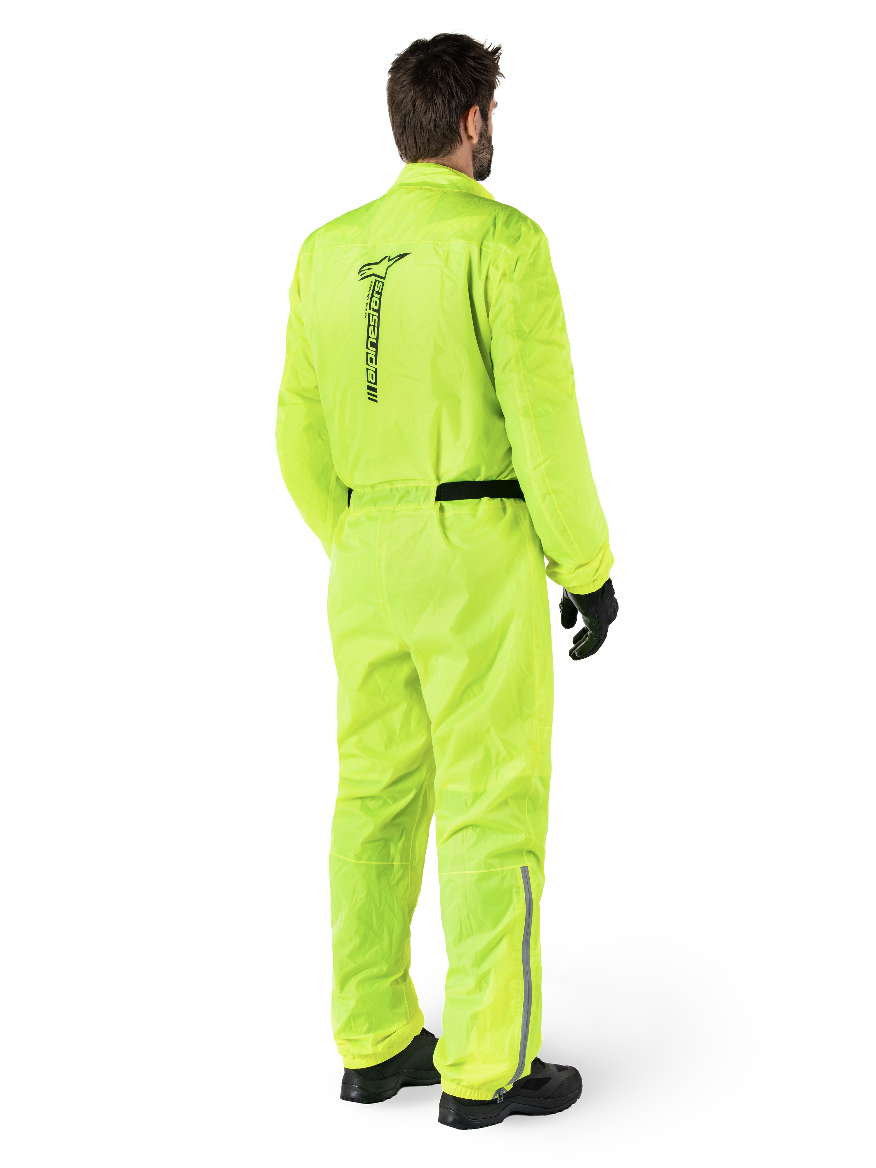 Alpinestars Hurricane Rain V2 Suit, Yellow Fluo, widok z tyłu motocyklisty w jednoczęściowym motocyklowym kombinezonie przeciwdeszczowym, z czarnym logo Alpinestars na plecach, czarnym paskiem w talii i odblaskowymi paskami na dolnej części nogawek, noszony z czarnymi rękawicami i butami