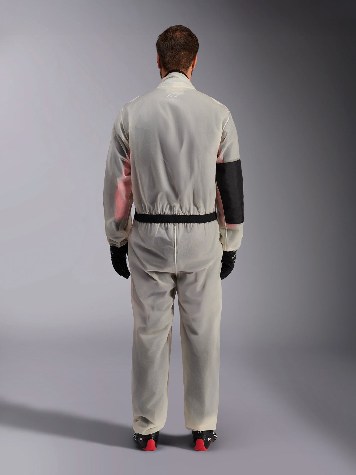 Alpinestars Kart Rain Suit, Karting Oversuit, Translucent Clear, back view showing a reinforced black panel on the right elbow for heat protection, elasticized waist with black trim, see-through material revealing a racing suit underneath, paired with black gloves and racing boots.