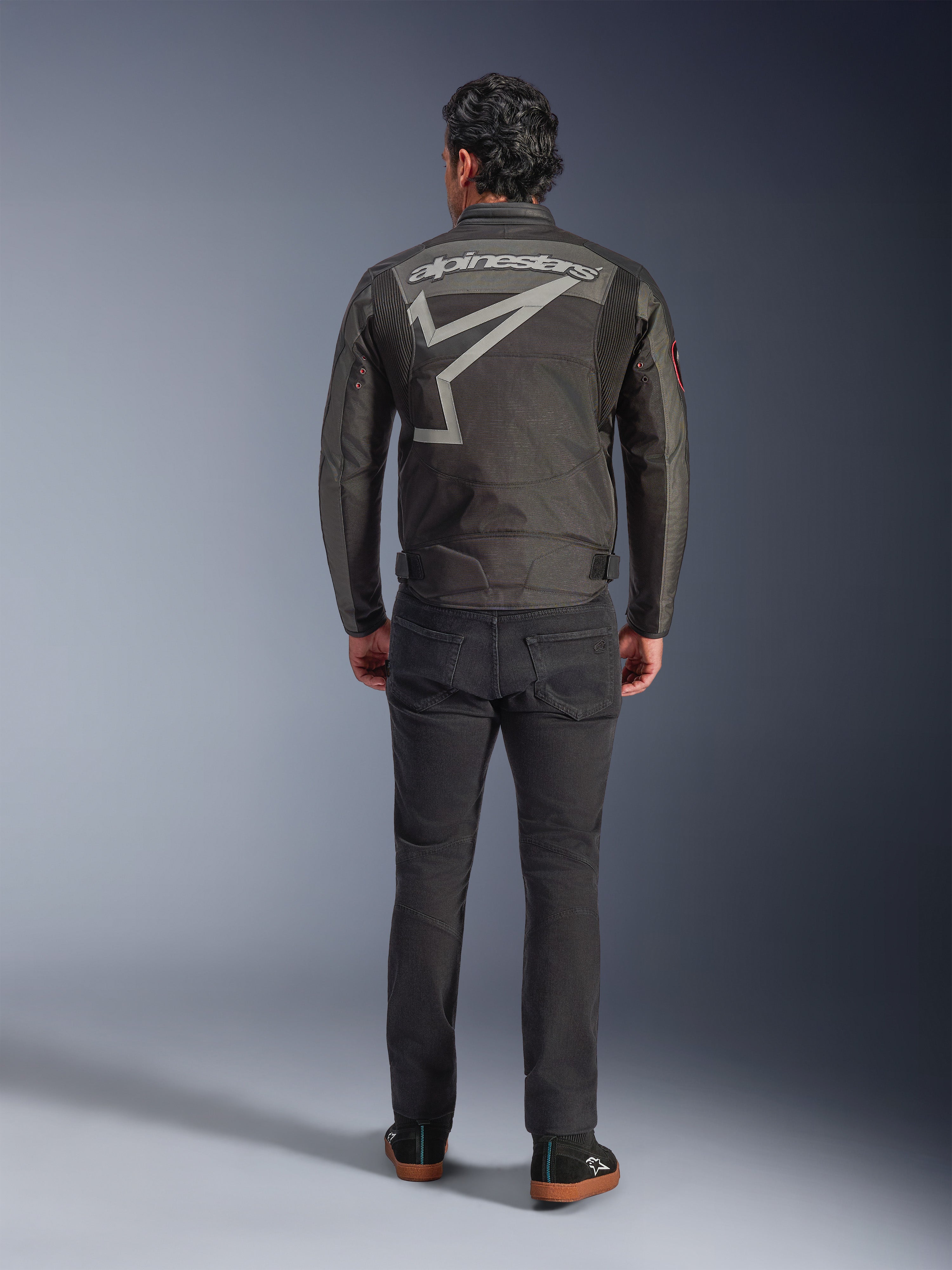 Alpinestars Axton Jacket, Motorcycle Jacket, Black Anthracite Red Tangerine, back view showing large grey Alpinestars logo and star graphic, accordion shoulder panels, worn with black pants and casual black shoes.