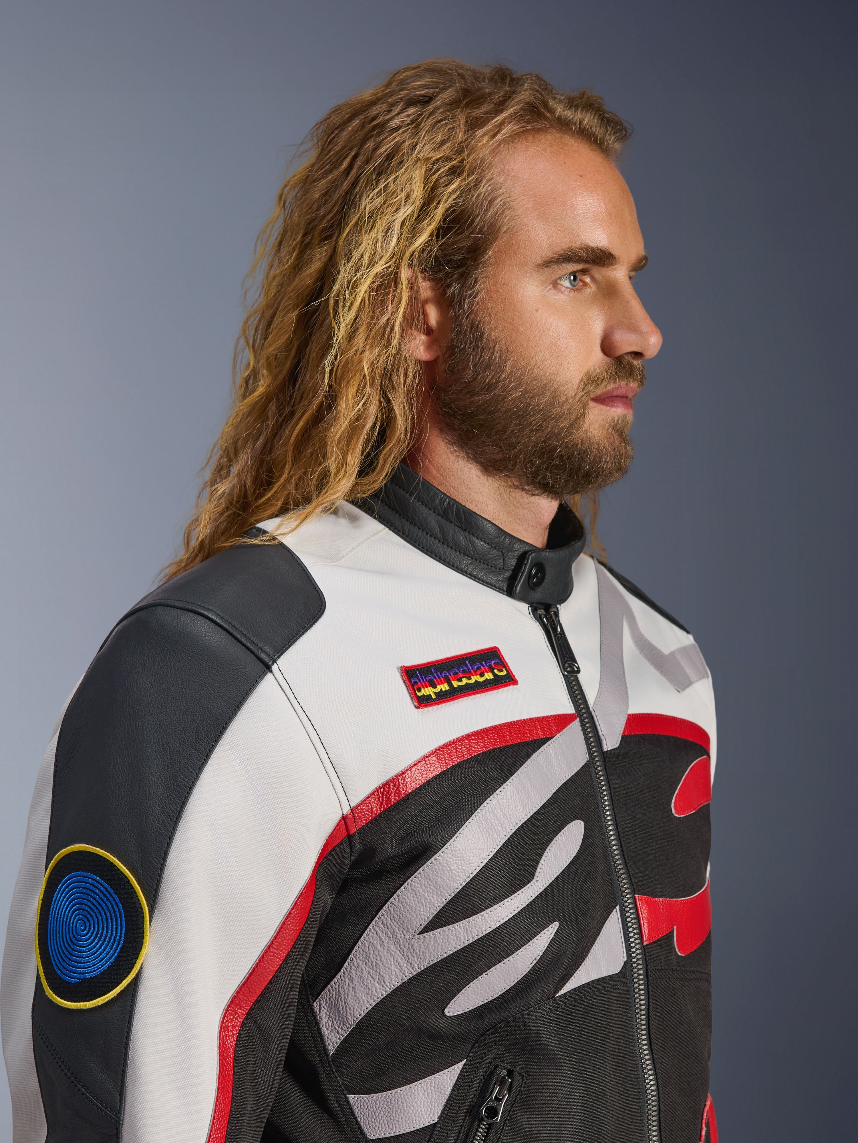 Man with long hair and beard wearing the Alpinestars Axton Jacket, Motorcycle Jacket, Black Off White and Bright Red, vintage racing-inspired design with branded patches and wrap-around logo, close-up profile view, urban contemporary style
