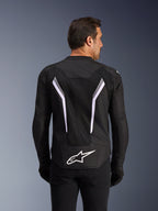 Alpinestars AST V3 Air Jacket, kurtka motocyklowa, czarna z białymi i ciemnoszarymi akcentami, widok z tyłu ukazujący obszerne siatkowe panele wentylacyjne, duże białe pasy w kształcie litery V oraz białe logo Alpinestars na dole pleców, noszona z czarnymi rękawicami i spodniami