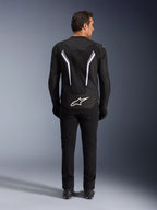 Kurtka Alpinestars AST V3 Air, widok z tyłu, czarna i ciemnoszara z białymi akcentami, duże białe logo Astar na dole, odblaskowe paski na plecach, siatkowa konstrukcja tekstylna, prezentowana z czarnymi spodniami i butami w sportowym stylu