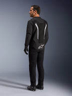 Alpinestars AST V3 Air Jacket, czarno-biała kurtka motocyklowa, widok z tyłu na modelu, z wstawkami z siatki zapewniającymi przepływ powietrza i białym logo na dole pleców, noszona z czarną techniczną odzieżą motocyklową.