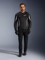 Męski model w kurtce Alpinestars AST V3 Air w kolorze czarnym z białymi akcentami, wyposażonej w panele wentylacyjne z siatki, o sportowym kroju, w zestawieniu z czarnymi technicznymi spodniami motocyklowymi, rękawicami i butami, widok z przodu, techniczna odzież motocyklowa do jazdy w ciepłe dni.