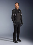 Kurtka Alpinestars AST V3 Air, czarna z białymi akcentami i logotypami, lekka kurtka motocyklowa z wentylacją siatkową, noszona przez mężczyznę z czarnymi spodniami, rękawicami i butami motocyklowymi, techniczna letnia odzież do jazdy sportowej i miejskiej.