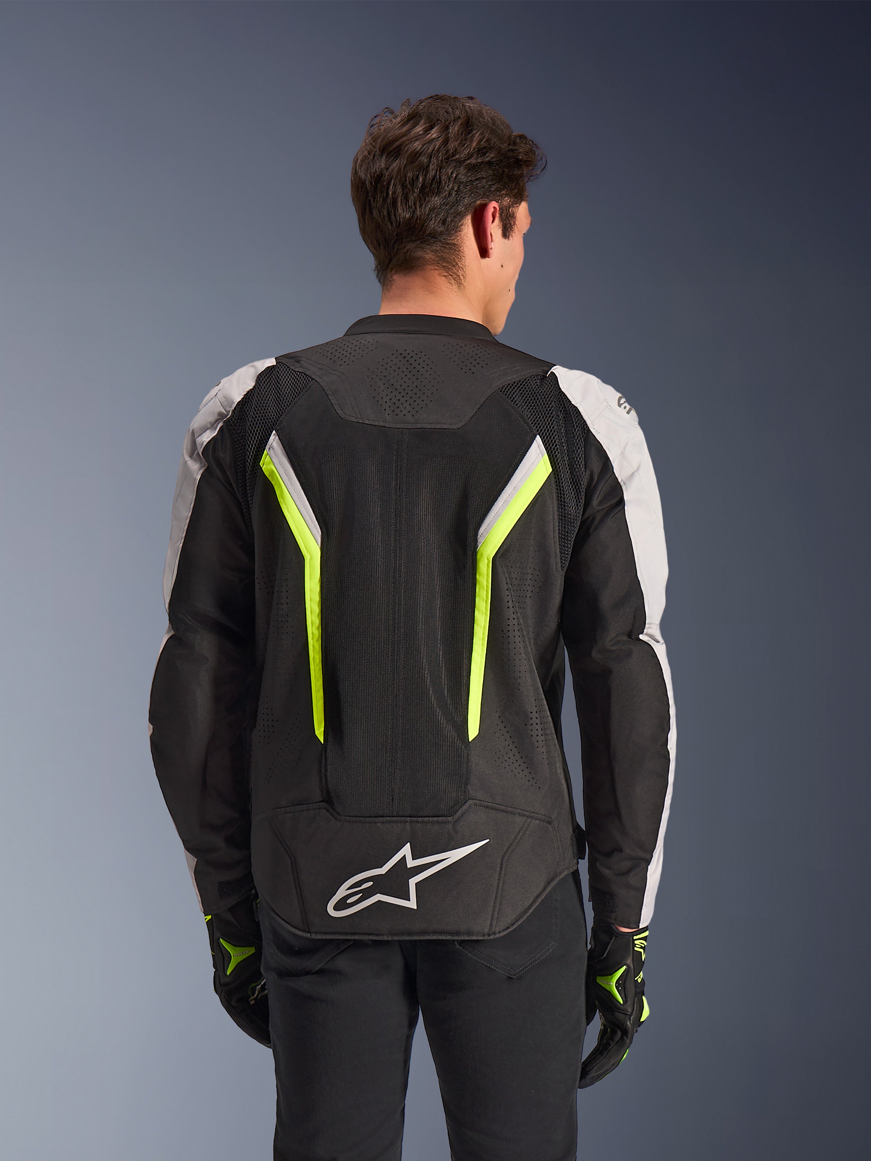 Alpinestars AST V3 Air Jacket, Black Silver Yellow Fluo, widok z tyłu na modelu, tekstylna kurtka motocyklowa z podwójnymi panelami wentylacyjnymi tech mesh, pionowymi neonowo-żółtymi pasami, srebrnymi rękawami i białym logo Alpinestars na dole pleców.