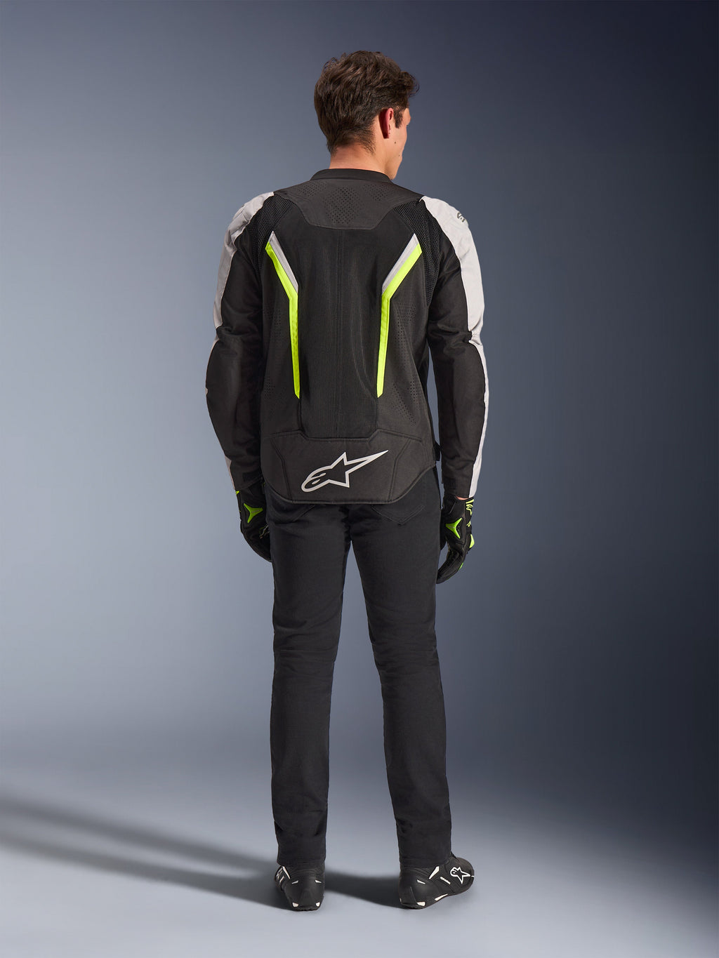 Alpinestars AST V3 Air Jacket, tekstylna kurtka motocyklowa, czarno-srebrna z żółtymi akcentami fluo, widok z tyłu ukazujący siatkowe panele wentylacyjne i fluorescencyjne akcenty, zestawiona z czarnymi spodniami i rękawicami, sportowa odzież motocyklowa