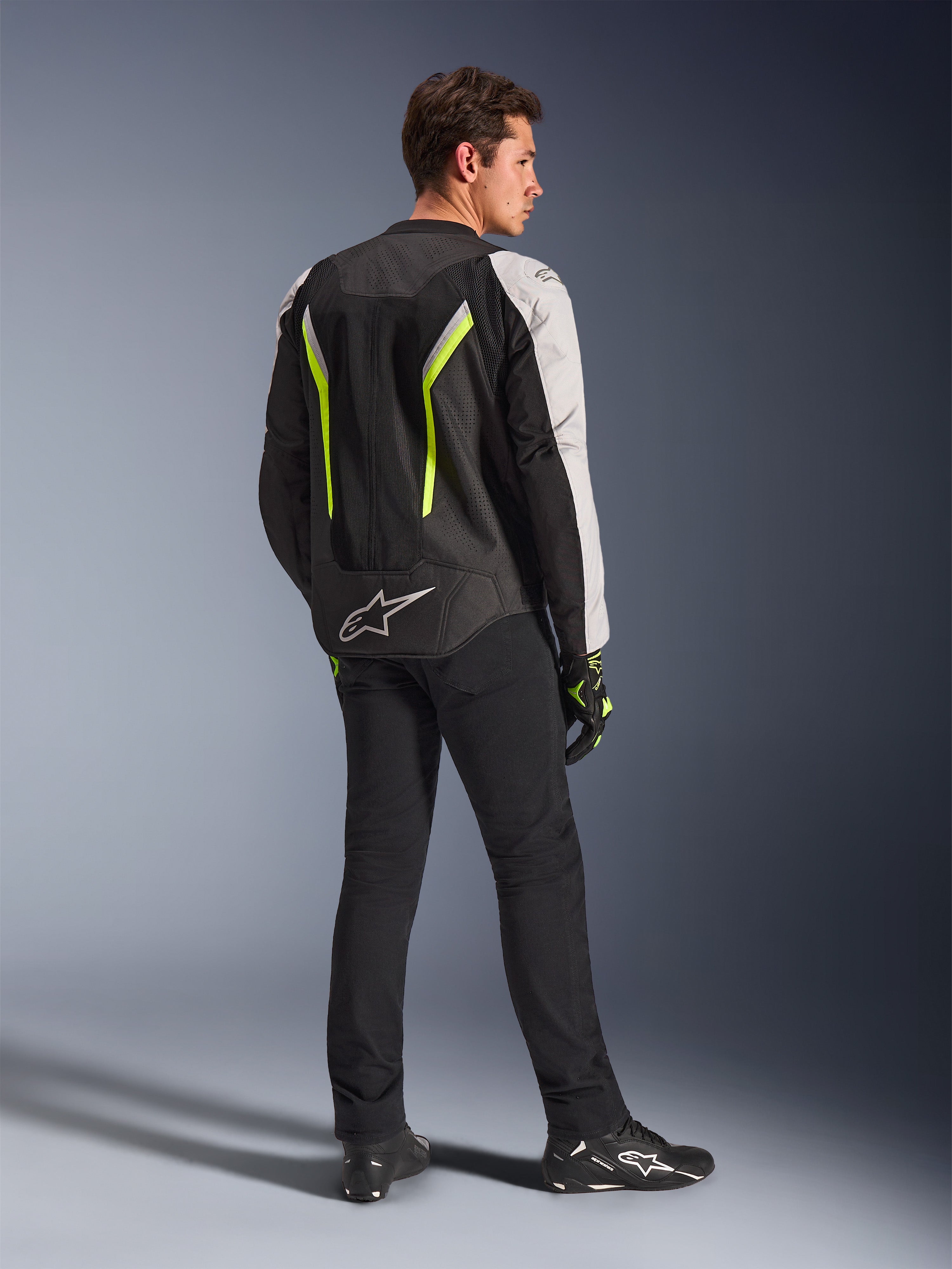 Alpinestars AST V3 Air Jacket w kolorze Black Silver Yellow Fluo, tekstylna kurtka motocyklowa widoczna od tyłu na motocykliście, z obszernymi panelami z siatki i fluorescencyjnymi żółtymi akcentami, zestawiona z czarnymi spodniami do jazdy, czarnymi rękawicami i czarnymi butami Alpinestars, prezentująca techniczny ubiór do jazdy sportowej