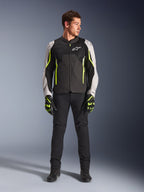 Męski model w kurtce Alpinestars AST V3 Air w kolorze Black Silver Yellow Fluo, z siatkowymi panelami wentylacyjnymi i fluorescencyjnymi akcentami, w zestawie z czarnymi spodniami motocyklowymi i pasującymi rękawicami, sportowa odzież wyczynowa.