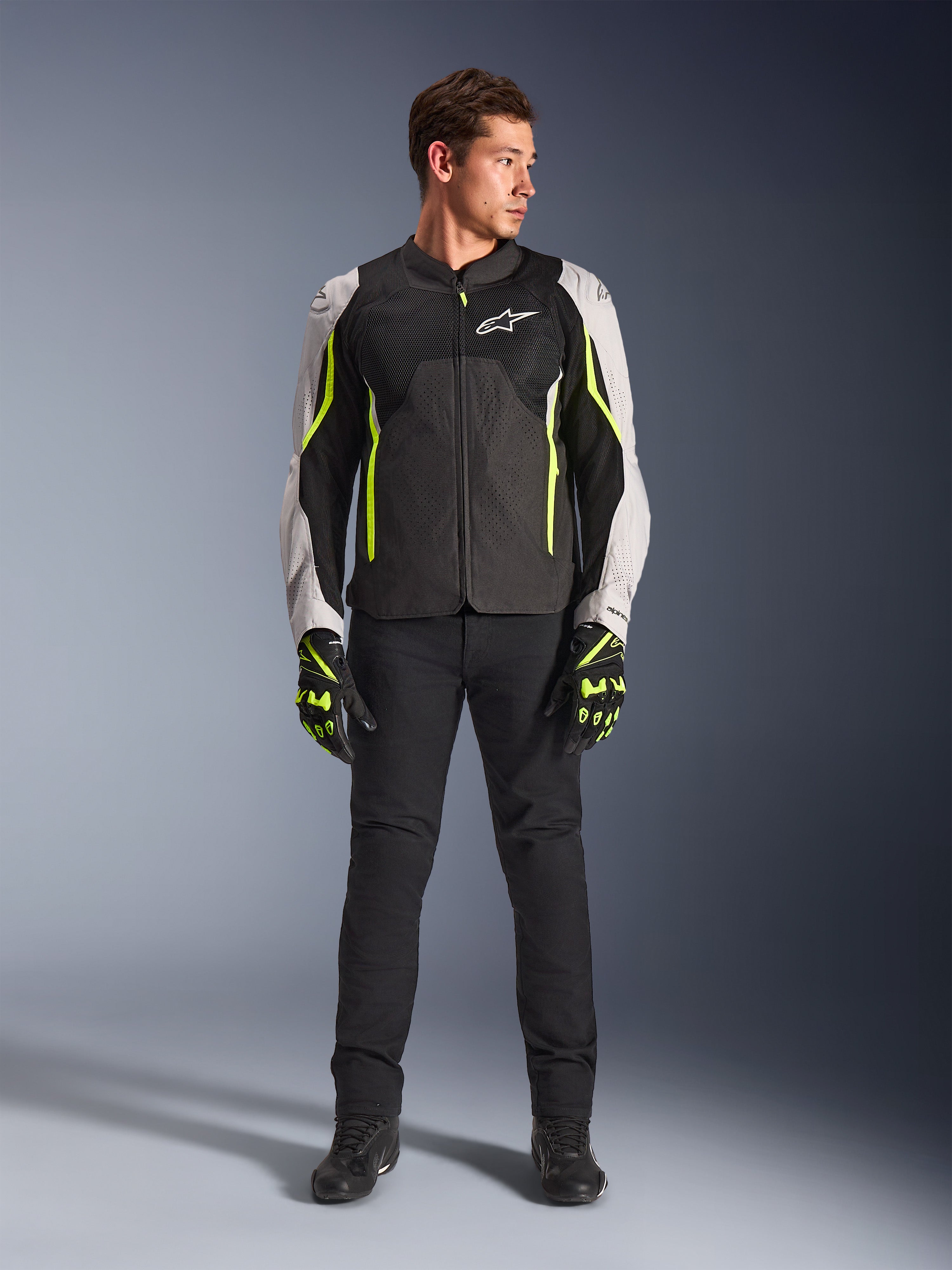 Męski model w kurtce Alpinestars AST V3 Air w kolorze Black Silver Yellow Fluo, z siatkowymi panelami wentylacyjnymi i fluorescencyjnymi akcentami, w zestawie z czarnymi spodniami motocyklowymi i pasującymi rękawicami, sportowa odzież wyczynowa.