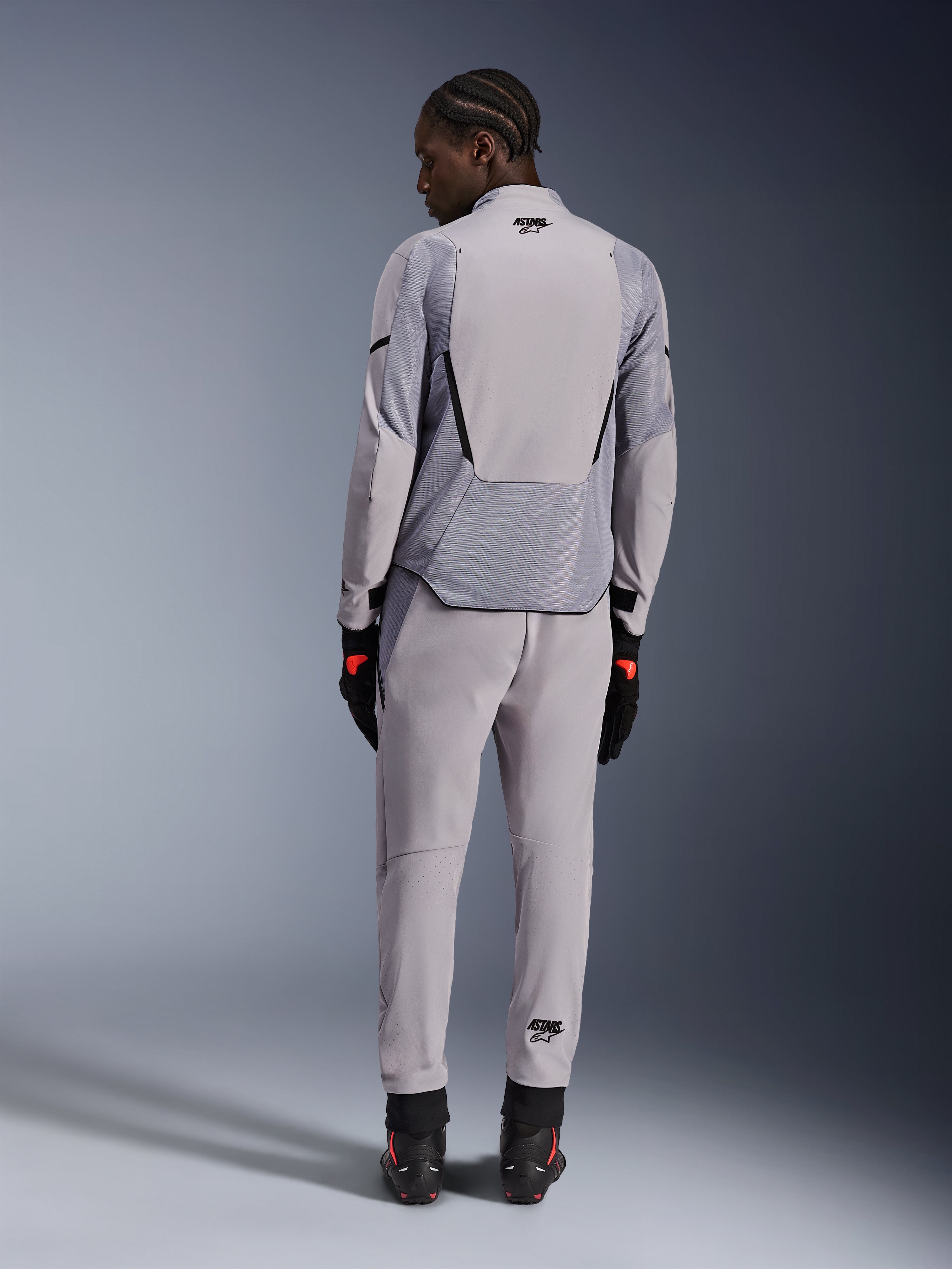 Kurtka Alpinestars Aeroshell Airflow, Mid Gray Anthracite, widok od tyłu na modelu, techniczne panele z siatki i laserowo perforowane strefy wentylacyjne, noszona z pasującymi szarymi spodniami i czarnym sprzętem motocyklowym, sportowy krój zapewniający wysoką wydajność.