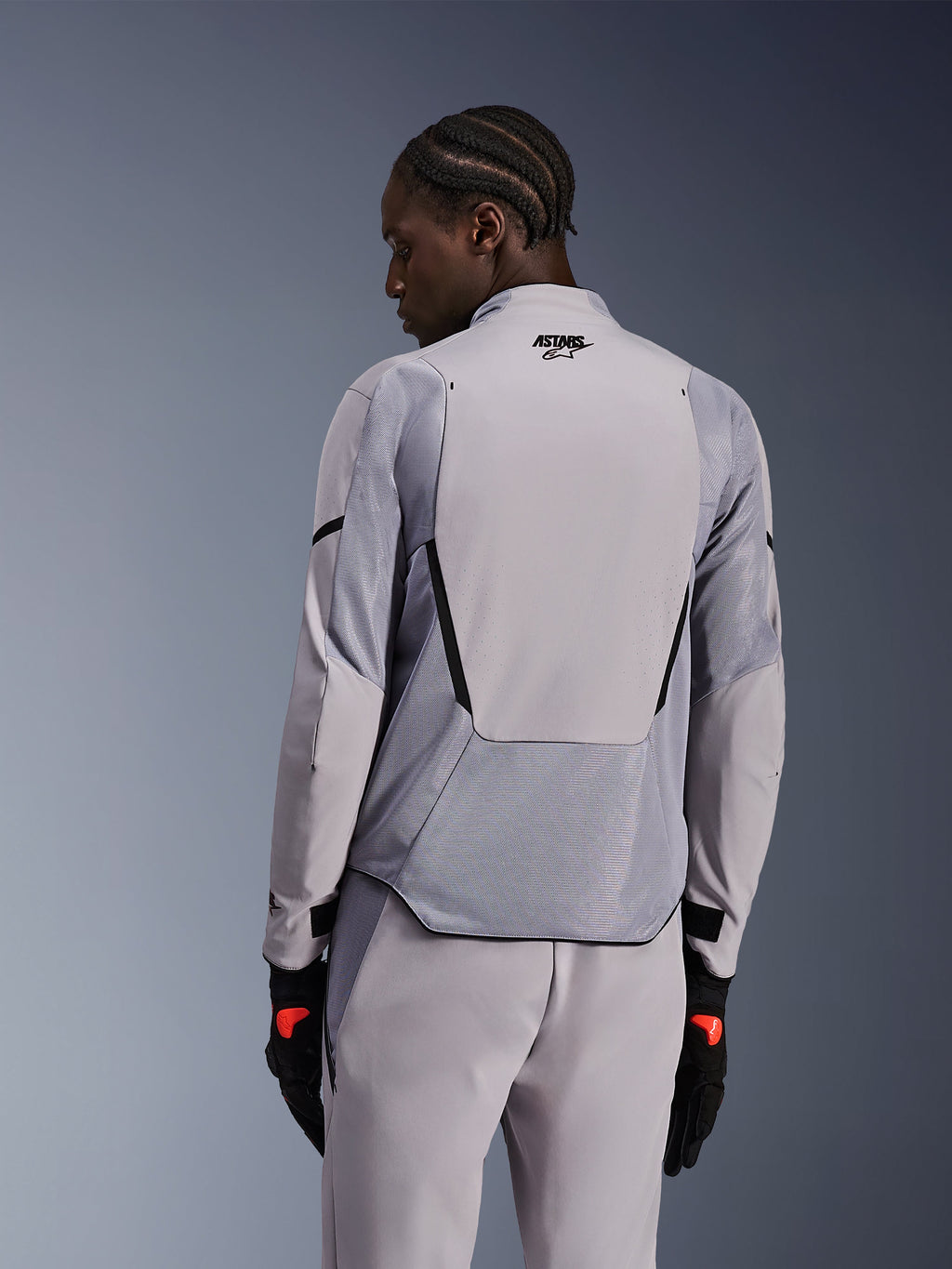 Kurtka Alpinestars Aeroshell Airflow w kolorze Mid Gray Anthracite, widok z tyłu prezentujący oddychające panele z siatki i perforacje laserowe, techniczna konstrukcja z elastycznego softshellu z wydłużonym panelem tylnym, sportowy styl z dopasowanymi szarymi spodniami i czarnymi rękawicami.
