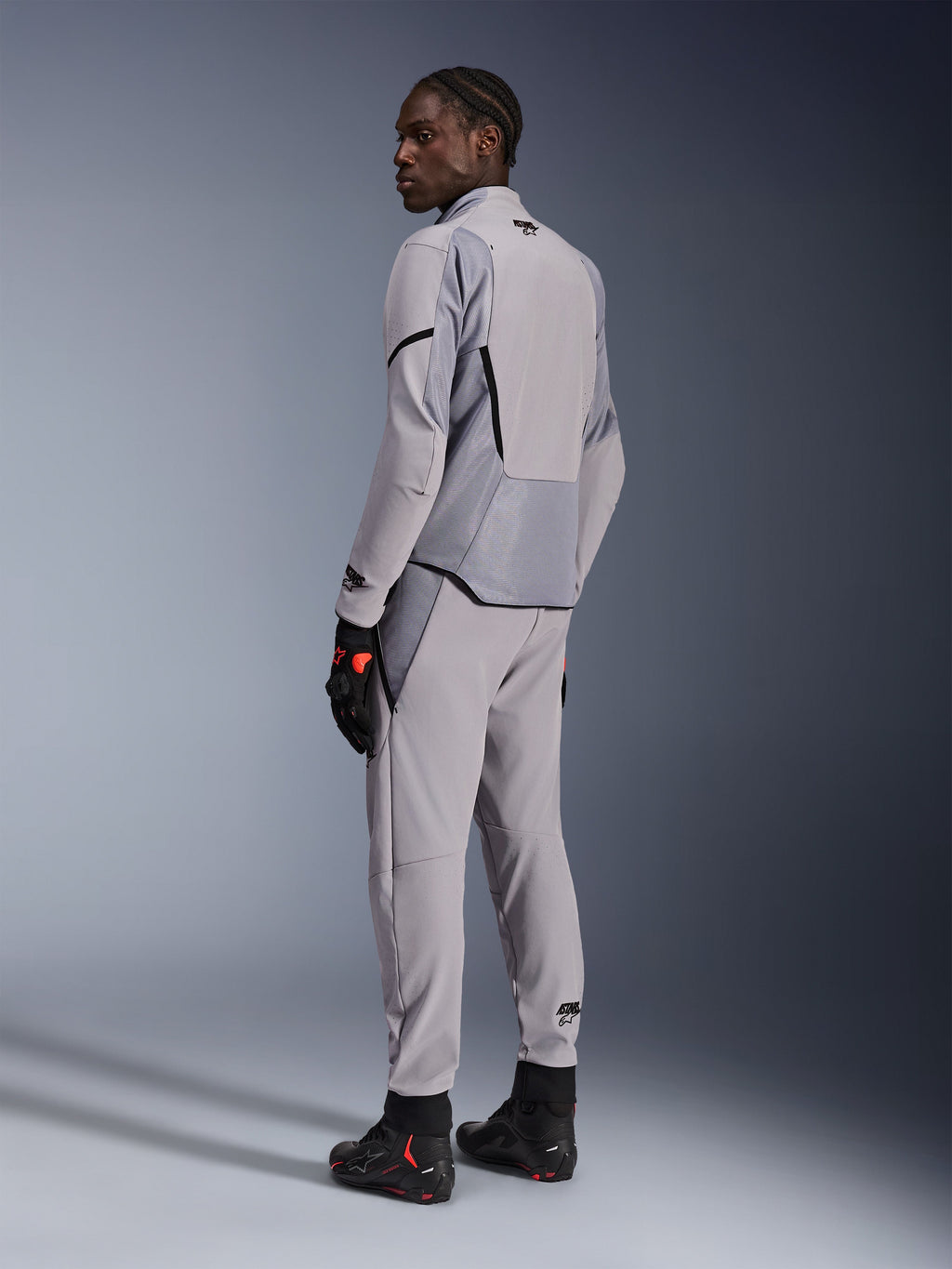 Kurtka Alpinestars Aeroshell Airflow w kolorze Mid Gray Anthracite, noszona z dopasowanymi spodniami i czarnymi rękawicami wyścigowymi, widok z tyłu ukazujący panele perforowane laserowo i sportowy krój, profesjonalna odzież motocyklowa
