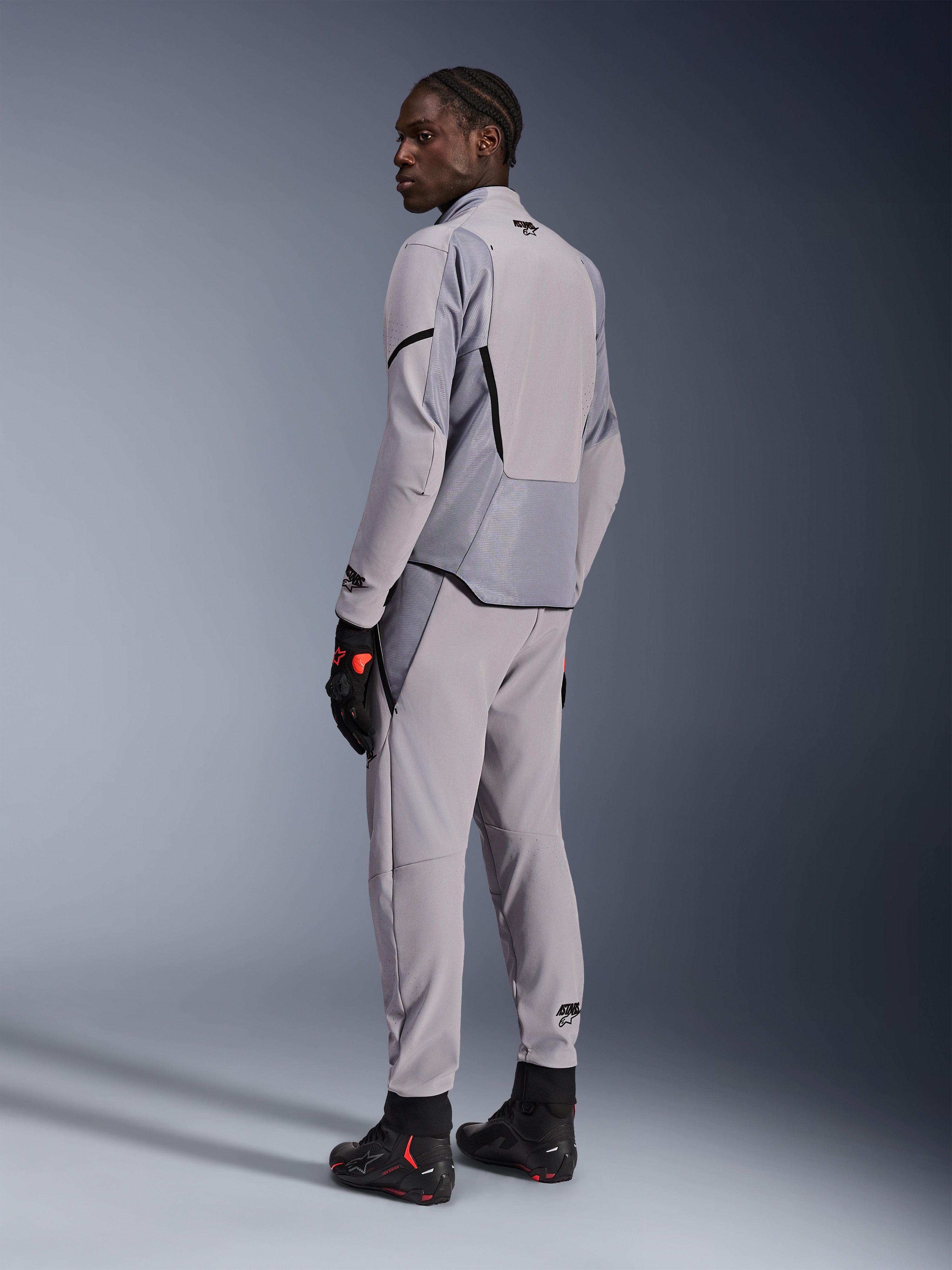 Kurtka Alpinestars Aeroshell Airflow w kolorze Mid Gray Anthracite, noszona z dopasowanymi spodniami i czarnymi rękawicami wyścigowymi, widok z tyłu ukazujący panele perforowane laserowo i sportowy krój, profesjonalna odzież motocyklowa