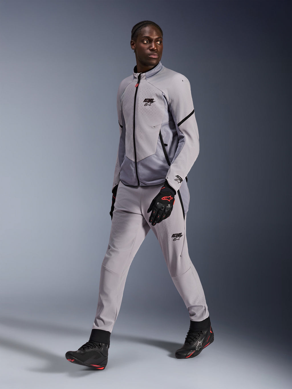 Kurtka Alpinestars Aeroshell Airflow w kolorze Mid Gray Anthracite, noszona przez męskiego modela z pasującymi szarymi spodniami motocyklowymi i czarnymi rękawicami, wyposażona w laserową perforację wentylacyjną na klatce piersiowej, o sportowym kroju.