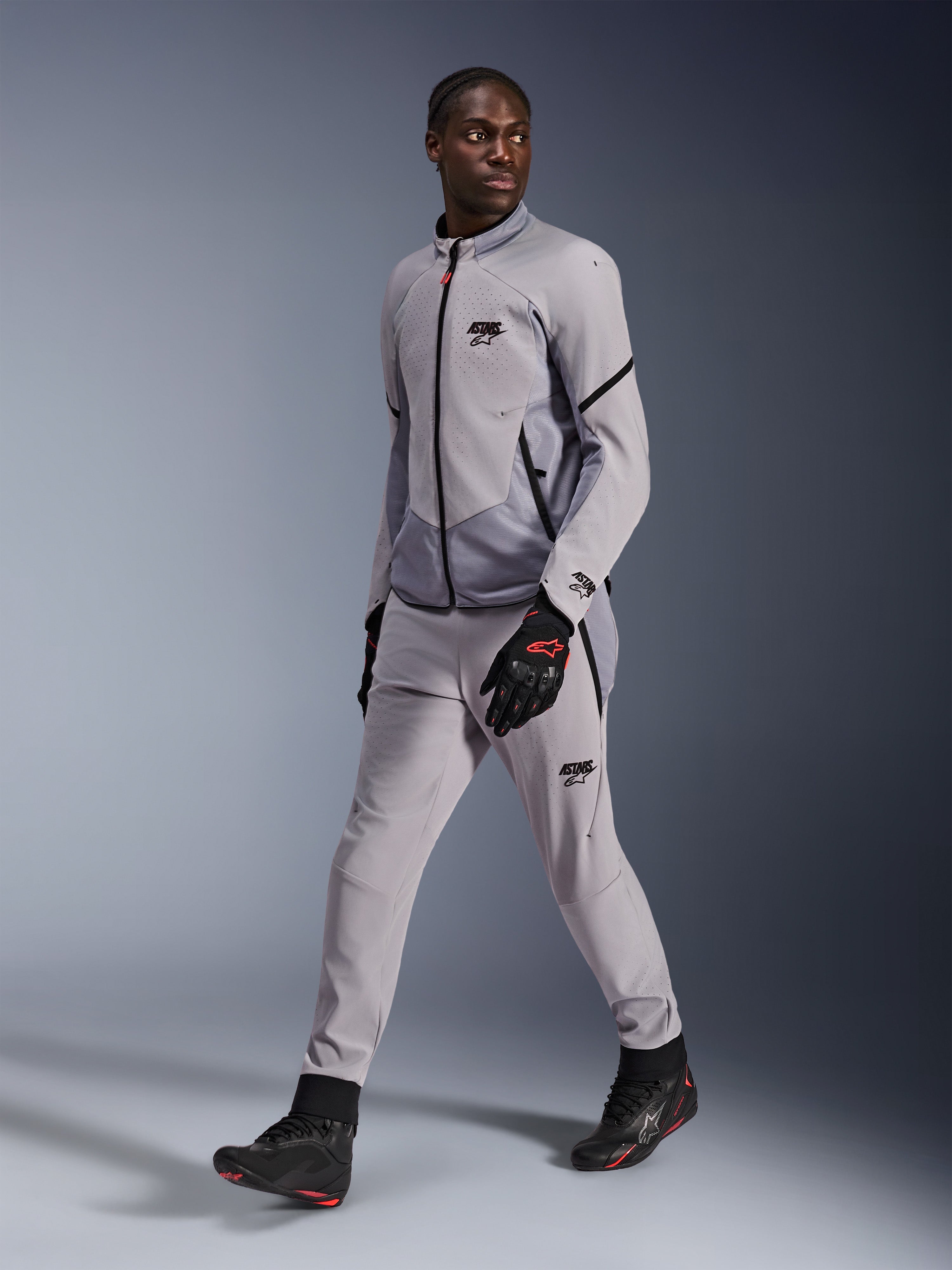 Kurtka Alpinestars Aeroshell Airflow w kolorze Mid Gray Anthracite, noszona przez męskiego modela z pasującymi szarymi spodniami motocyklowymi i czarnymi rękawicami, wyposażona w laserową perforację wentylacyjną na klatce piersiowej, o sportowym kroju.