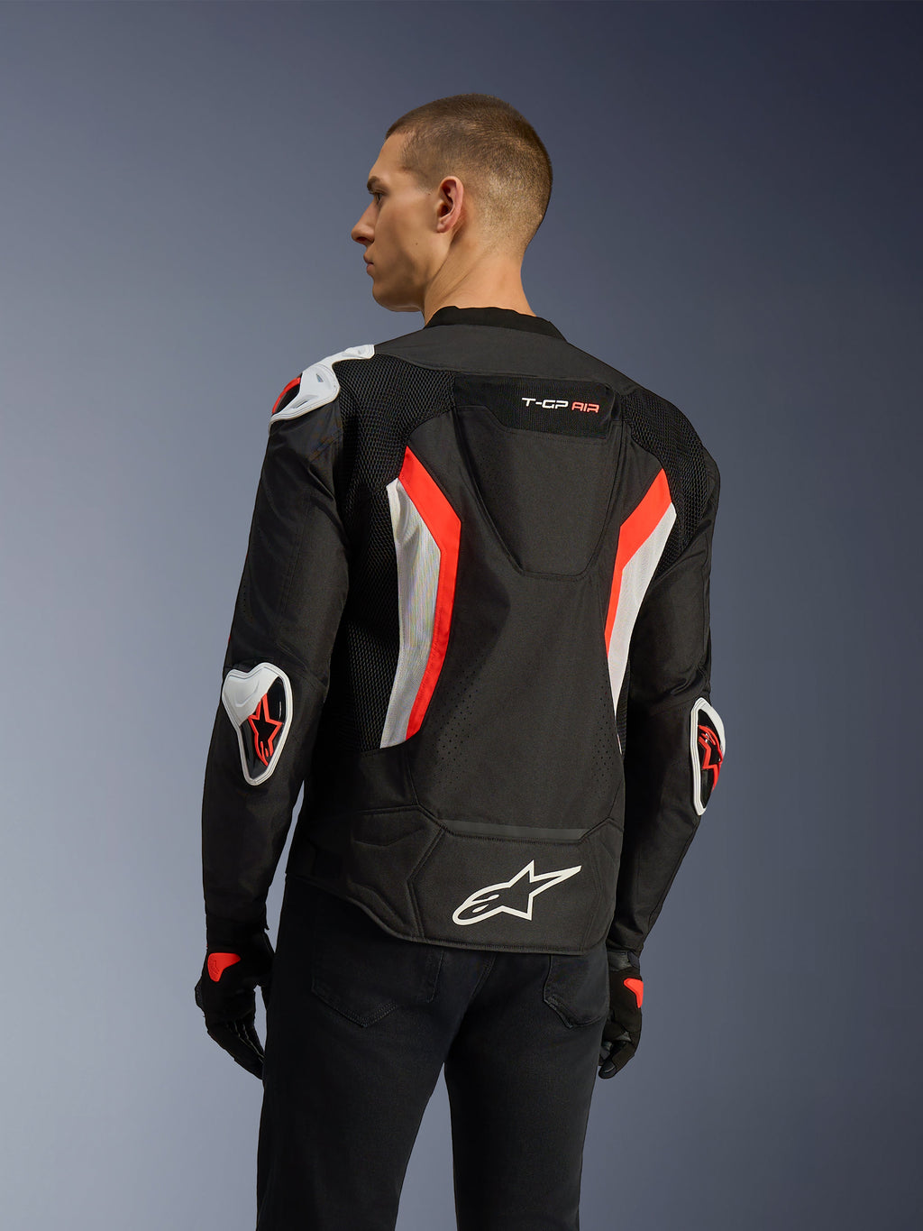 Kurtka Alpinestars T-GP Air w kolorze Black Red Fluo, widok z tyłu pod kątem trzech czwartych na modelu, podkreślający garb aerodynamiczny, zewnętrzne ochraniacze Racing GP DFS na ramionach i łokciach, laserowo perforowane panele mesh oraz białe i czerwone akcenty fluo, tekstylna kurtka motocyklowa o sportowym kroju.