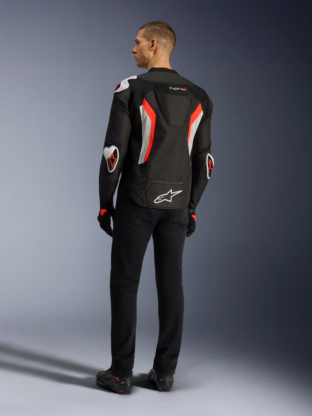 Kurtka Alpinestars T-GP Air, kurtka motocyklowa w kolorystyce Black Red Fluo, widok z tyłu na modelu, z panelami wentylacyjnymi z siatki, garbem aerodynamicznym i białymi zewnętrznymi ochraniaczami na łokciach i ramionach, noszona z czarnymi spodniami i butami motocyklowymi