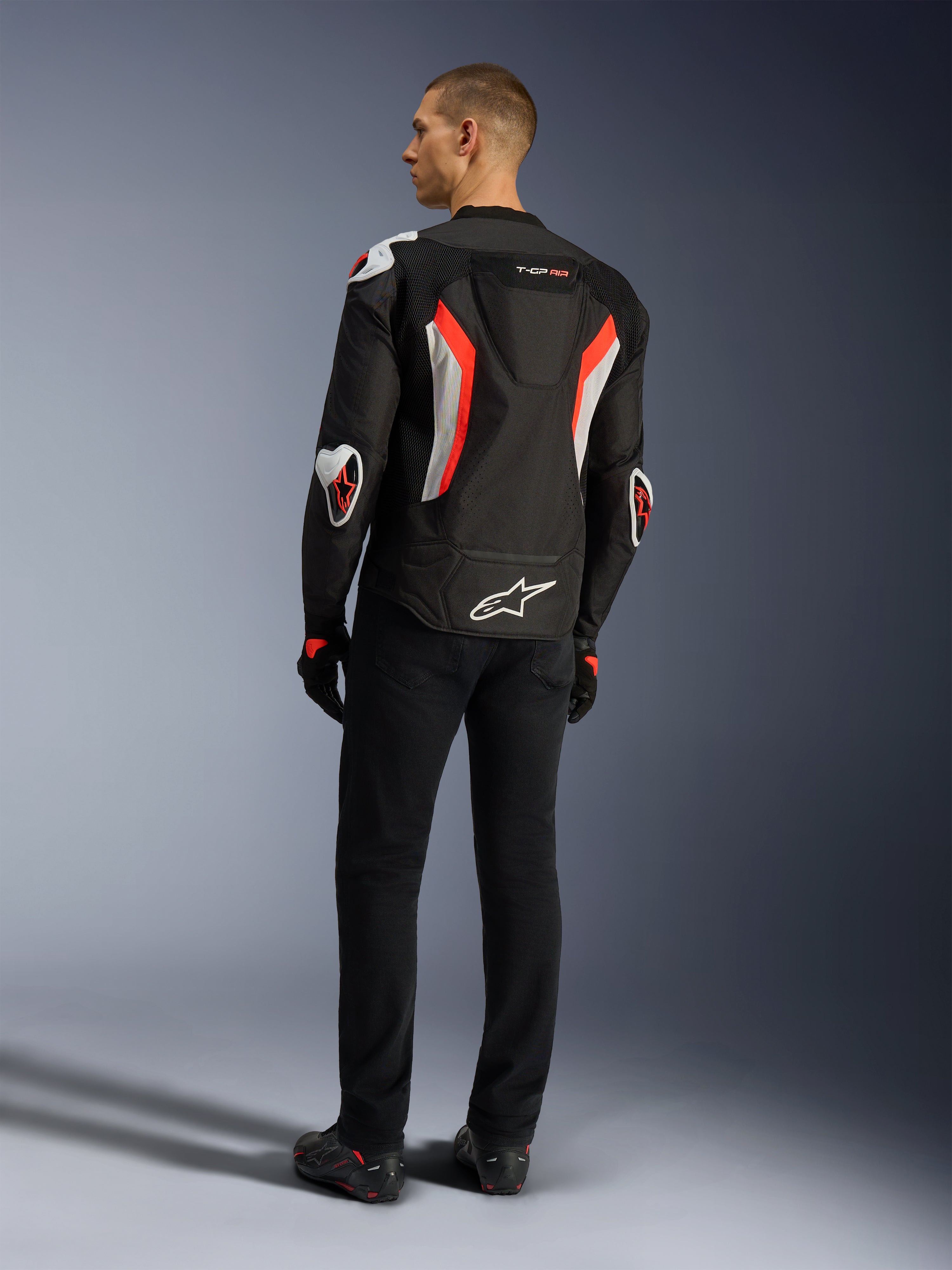 Kurtka Alpinestars T-GP Air, kurtka motocyklowa w kolorystyce Black Red Fluo, widok z tyłu na modelu, z panelami wentylacyjnymi z siatki, garbem aerodynamicznym i białymi zewnętrznymi ochraniaczami na łokciach i ramionach, noszona z czarnymi spodniami i butami motocyklowymi