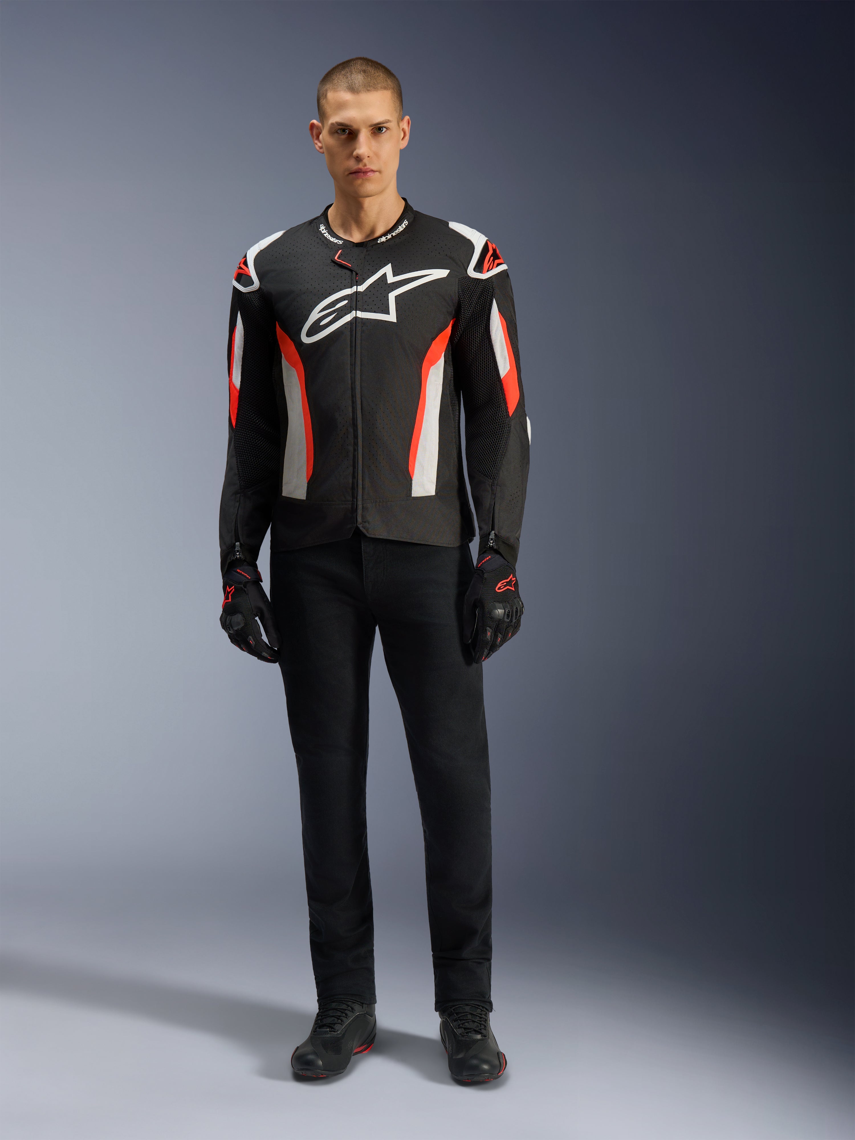 Model męski w kurtce Alpinestars T-GP Air w kolorze Black Red Fluo, tekstylna kurtka motocyklowa z białymi i jaskrawoczerwonymi akcentami, duże białe logo Alpinestars na piersi, panele wentylacyjne z siatki, czarne spodnie techniczne i rękawice motocyklowe, sportowy krój