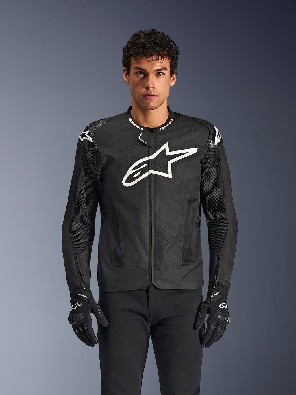 Alpinestars T-GP Air Jacket, Black textile motorcycle jacket with a large white star logo on the chest, features perforated ventilation panels and 3D mesh, external shoulder sliders, worn with black and white gloves, sleek sporty design for warm-weather road riding