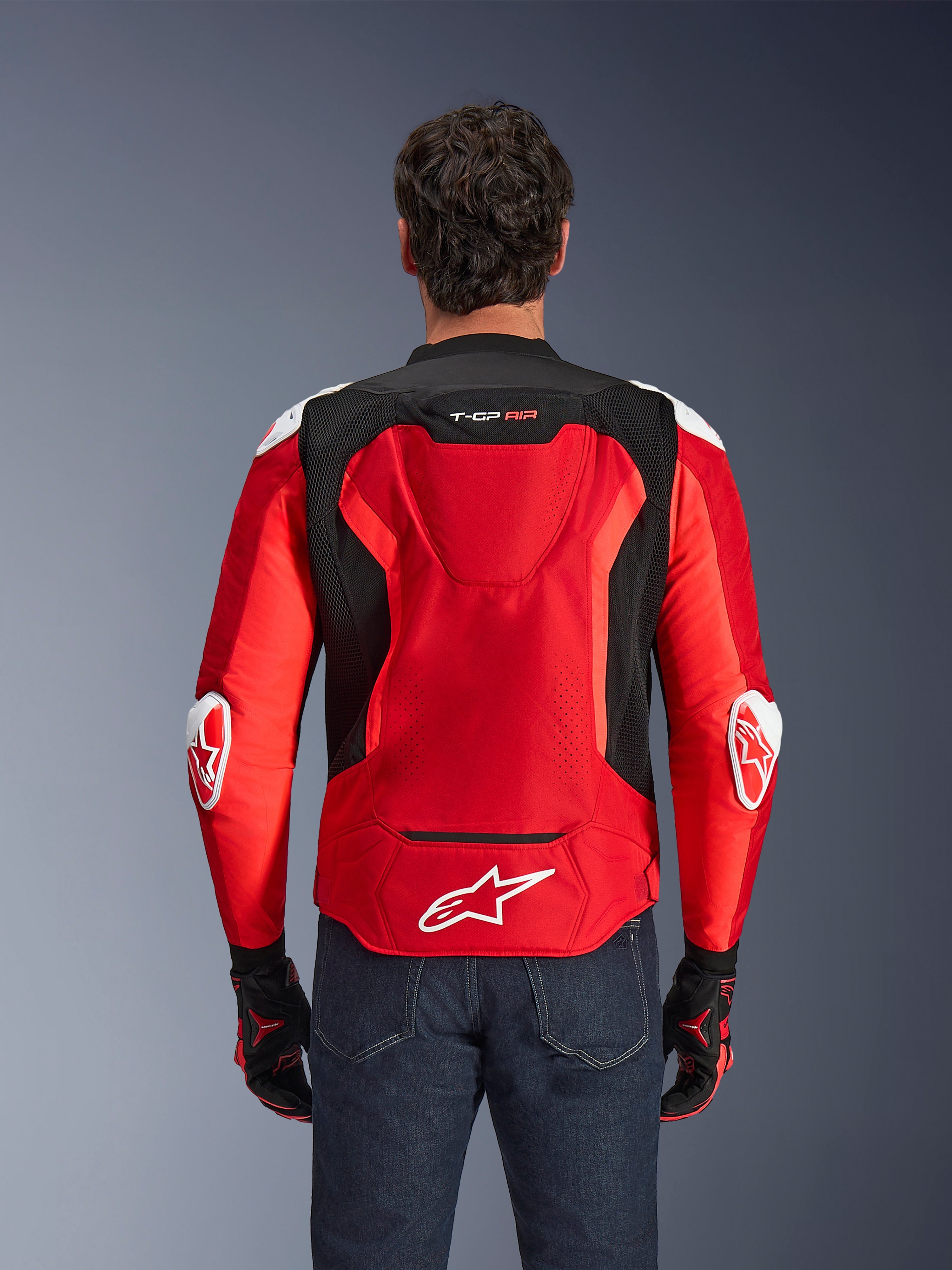 Kurtka Alpinestars T-GP Air, tekstylna kurtka motocyklowa, jaskrawoczerwona i czarna z białymi akcentami, widok z tyłu ukazujący garb aerodynamiczny i siatkowe panele wentylacyjne, z logo gwiazdy Alpinestars i brandingiem T-GP Air, noszona z ciemnymi dżinsami i rękawicami wyścigowymi.
