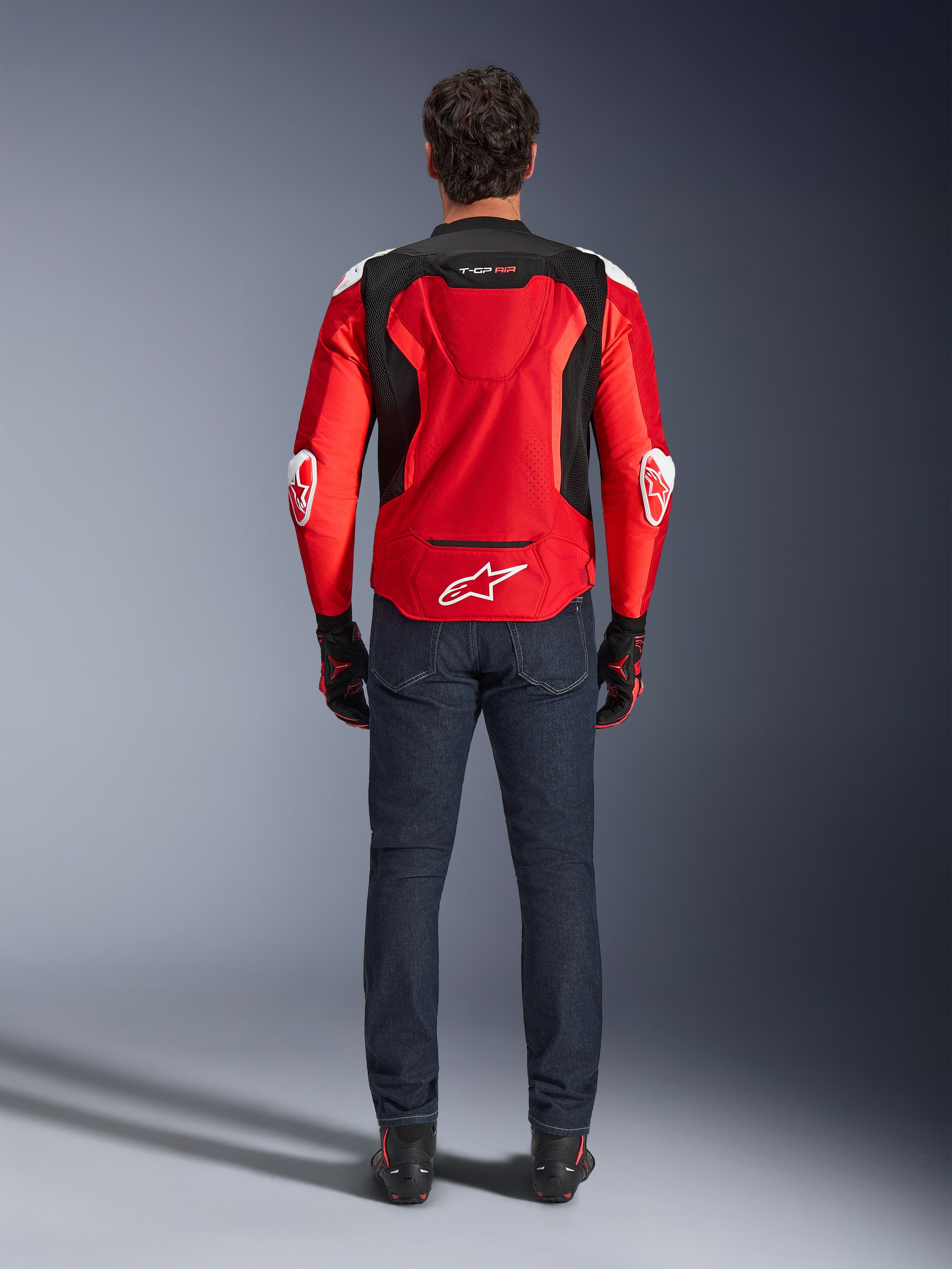 Kurtka Alpinestars T-GP Air, widok z tyłu, jaskrawoczerwona i czarna, z białym logo w dolnej części pleców i oznaczeniem „T-GP AIR”, noszona z ciemnoniebieskimi dżinsami i czarno-czerwonymi rękawicami, sportowa odzież motocyklowa