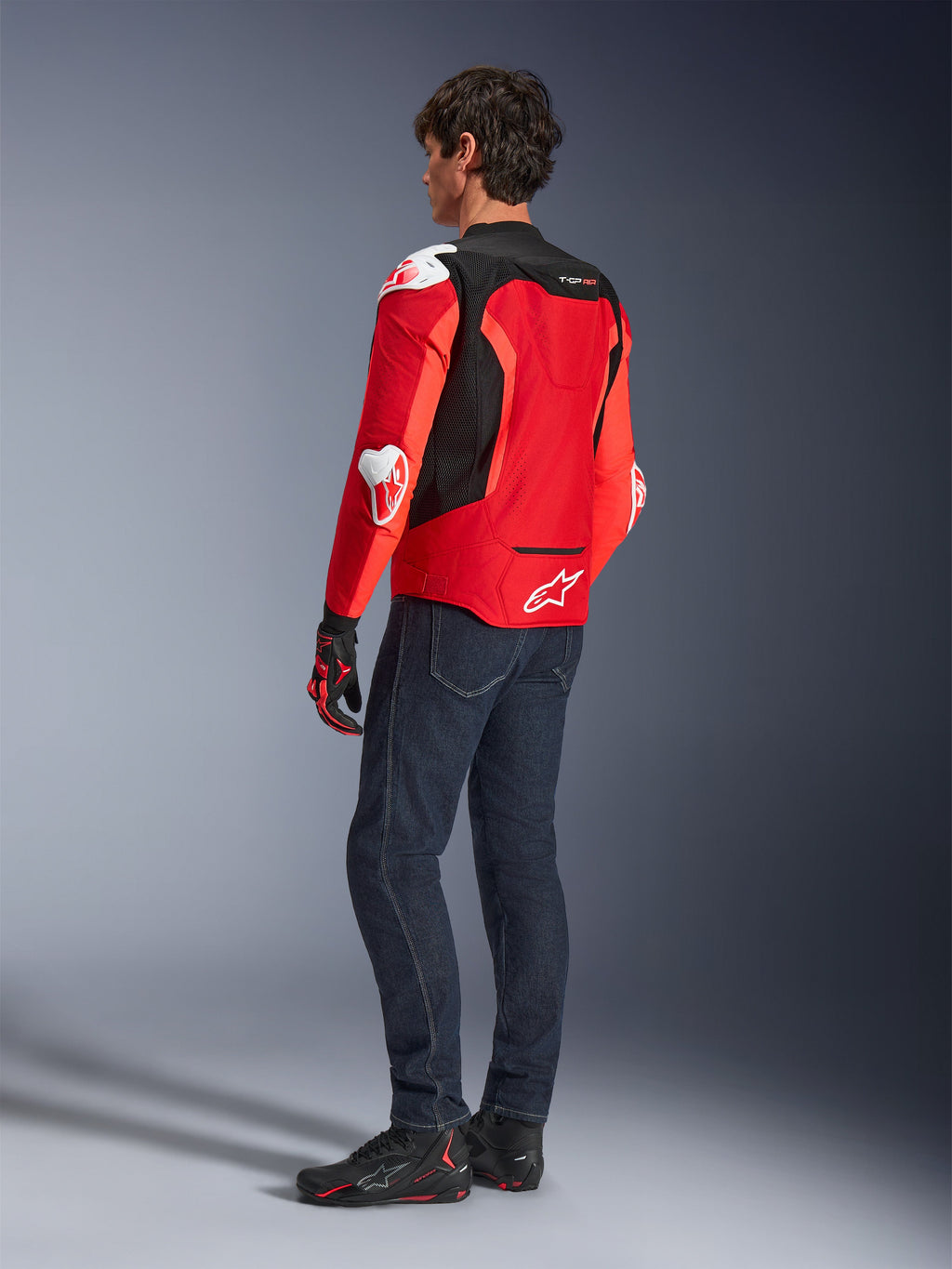 Kurtka Alpinestars T-GP Air, widok z tyłu na mężczyznę ubranego w jaskrawoczerwoną i neonowo-czerwoną kurtkę tekstylną z czarnymi panelami z siatki i białymi sliderami, w zestawieniu z ciemnoniebieskimi dżinsami i czarnymi butami motocyklowymi, sportowa odzież motocyklowa