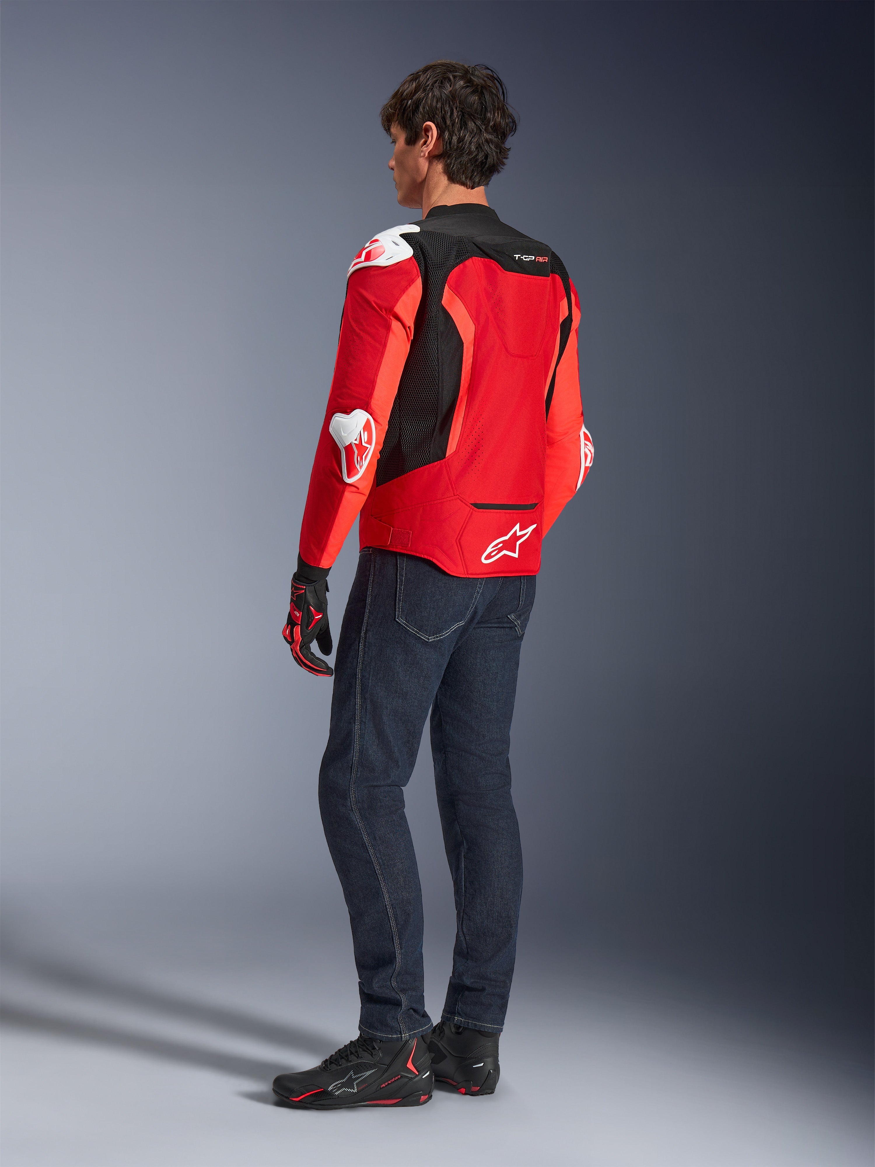 Kurtka Alpinestars T-GP Air, widok z tyłu na mężczyznę ubranego w jaskrawoczerwoną i neonowo-czerwoną kurtkę tekstylną z czarnymi panelami z siatki i białymi sliderami, w zestawieniu z ciemnoniebieskimi dżinsami i czarnymi butami motocyklowymi, sportowa odzież motocyklowa